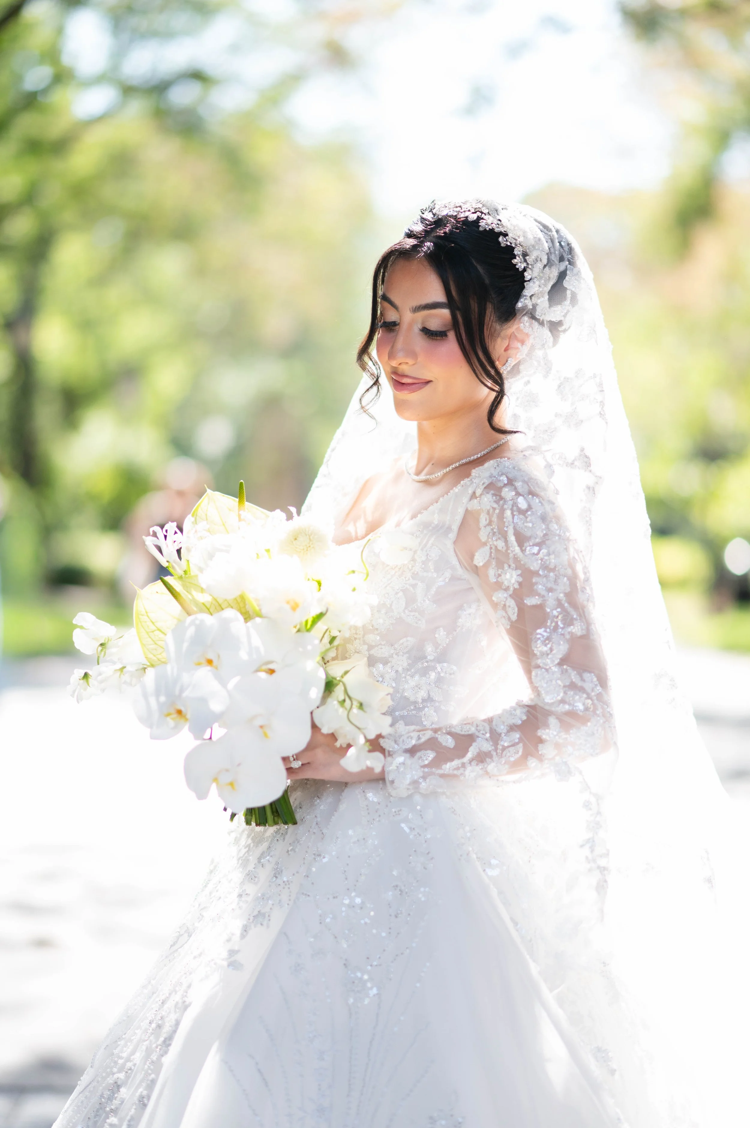 A bride in a lace wedding dress and veil holding a bouquet of white flowers outdoors on a sunny day.