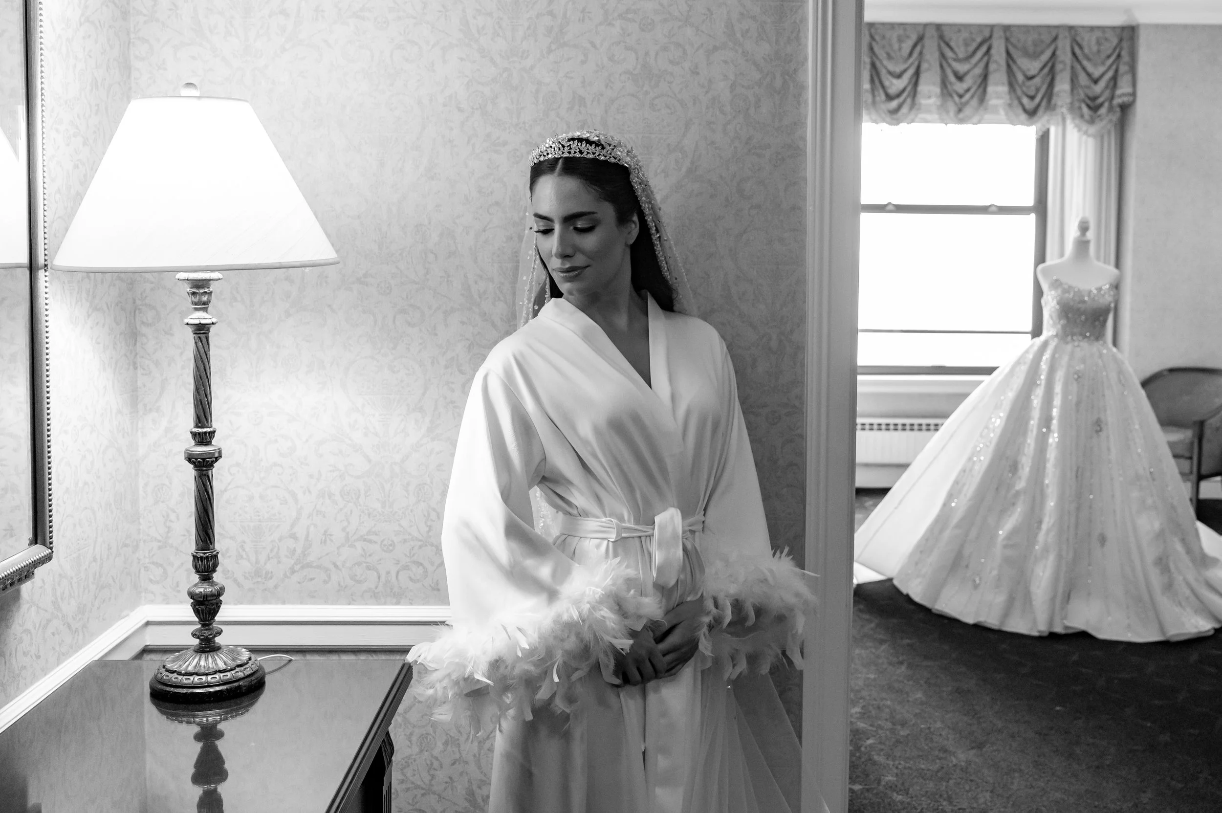 A bride in a satin robe with feather trim stands in a room, looking down. A wedding gown with a full skirt and beaded details is displayed on a mannequin near the window.