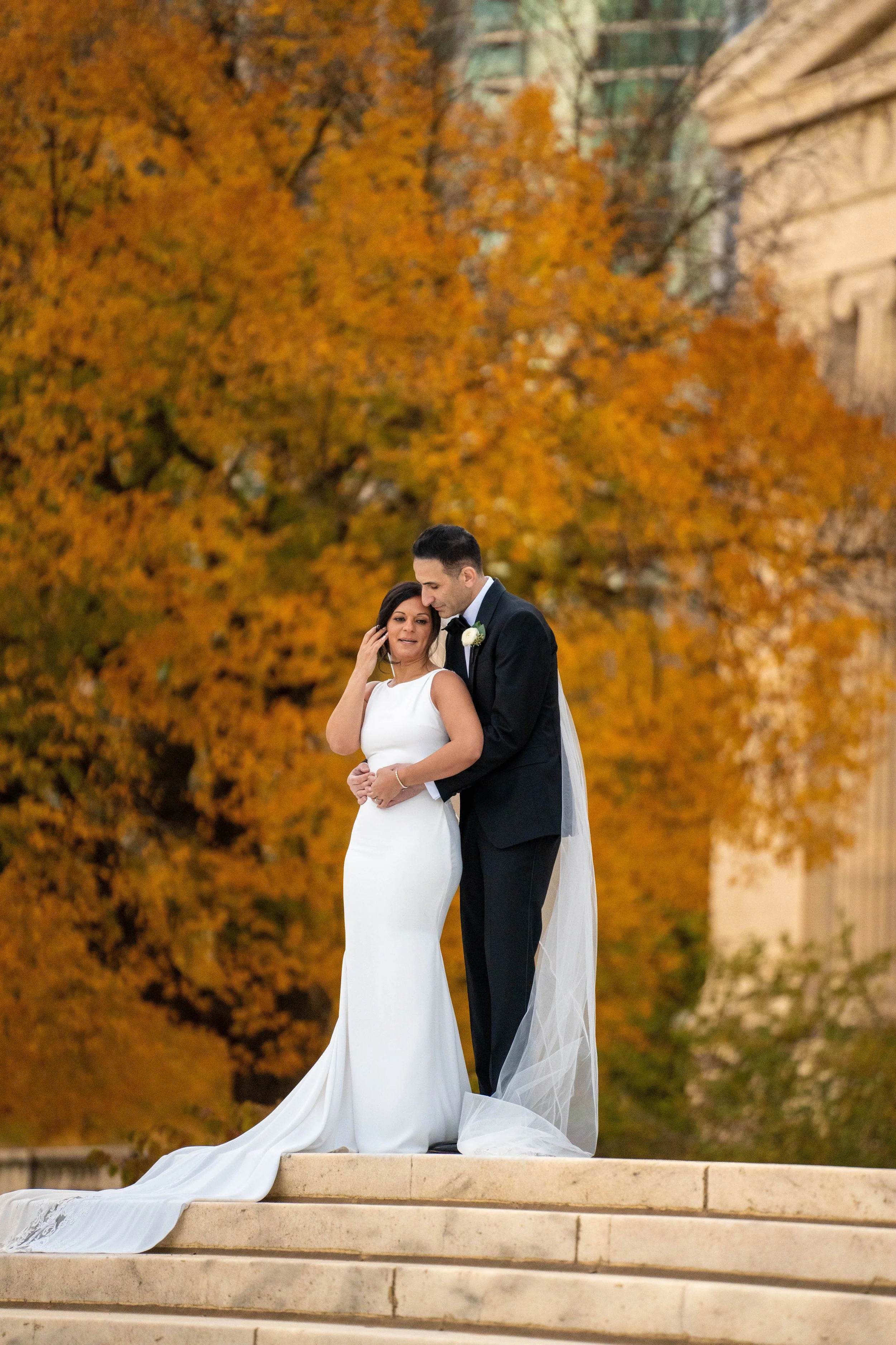 A bride and groom standing on steps outdoors during fall with orange trees and a building in the background