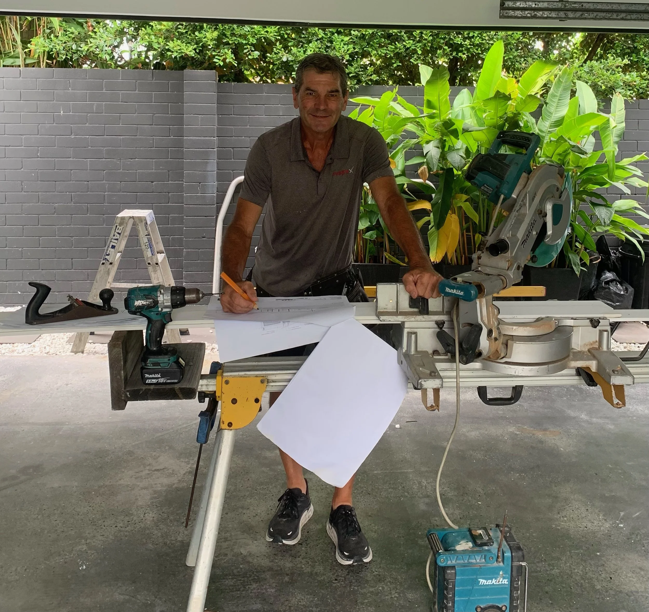 A man in a gray polo shirt and black sneakers stands at a workbench with tools, including a power drill and a miter saw, in an outdoor space with a gray brick wall and lush green plants in the background.