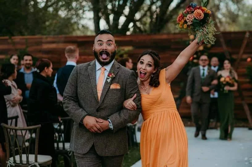 A happy woman in an orange dress holding a bouquet celebrates at an outdoor wedding reception.