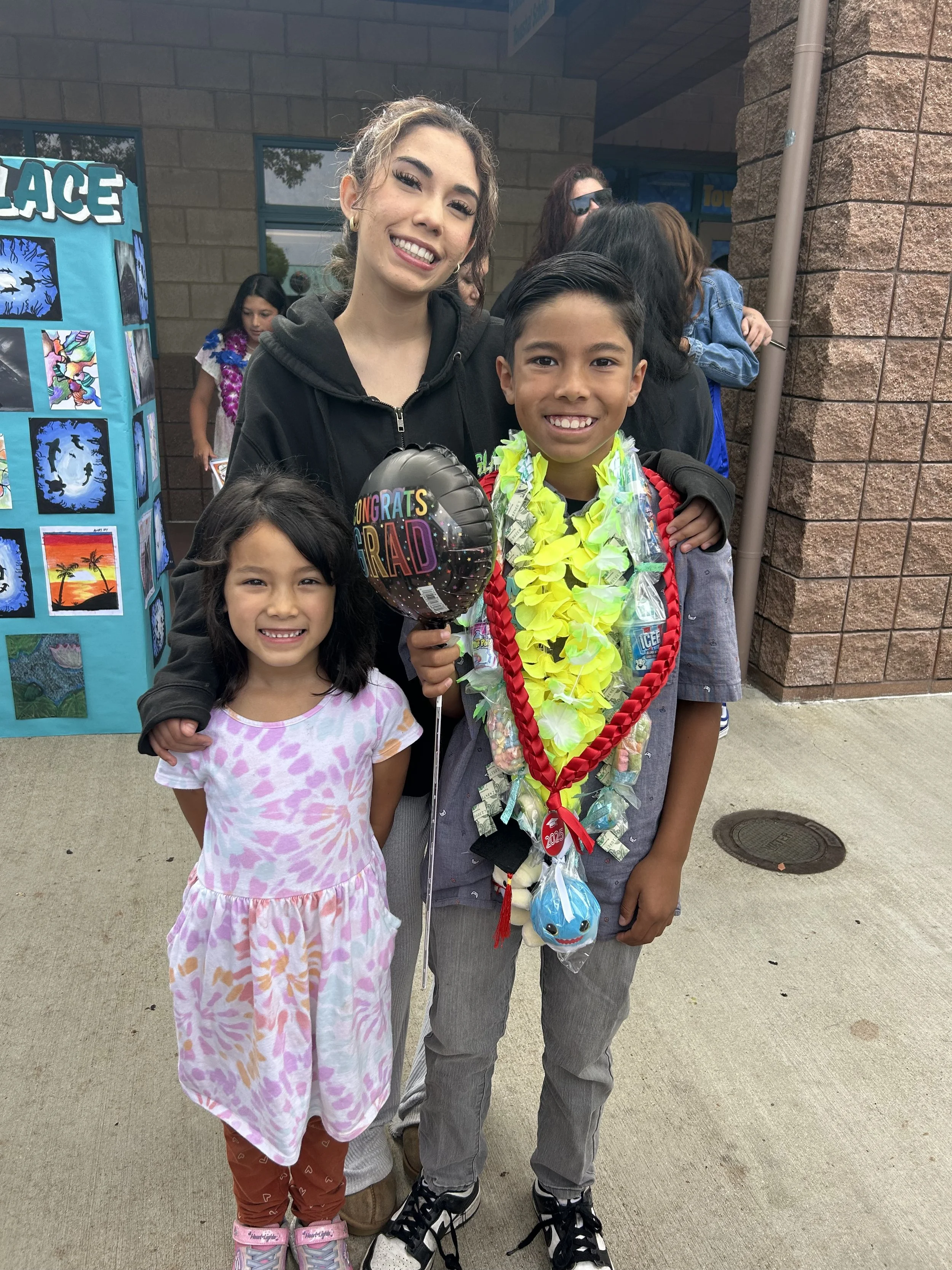 A group of three children and an adult woman standing outside, celebrating a graduation. The boy in the center is wearing a flower lei and holding a congratulatory balloon. The girl on the left is wearing a colorful dress. The woman behind them is smiling, and there are other people and a display board in the background.