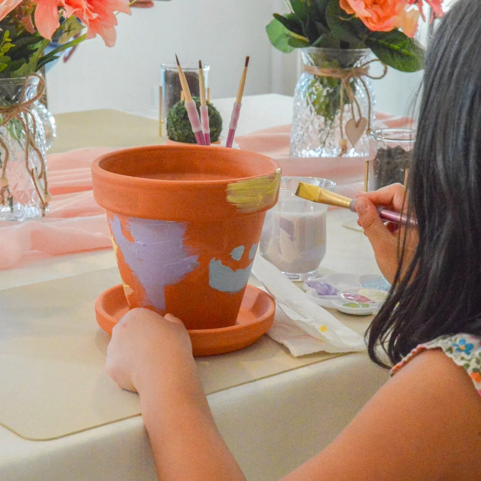 A young girl painting a flowerpot with a paintbrush at a decorated table with flowers and art supplies.