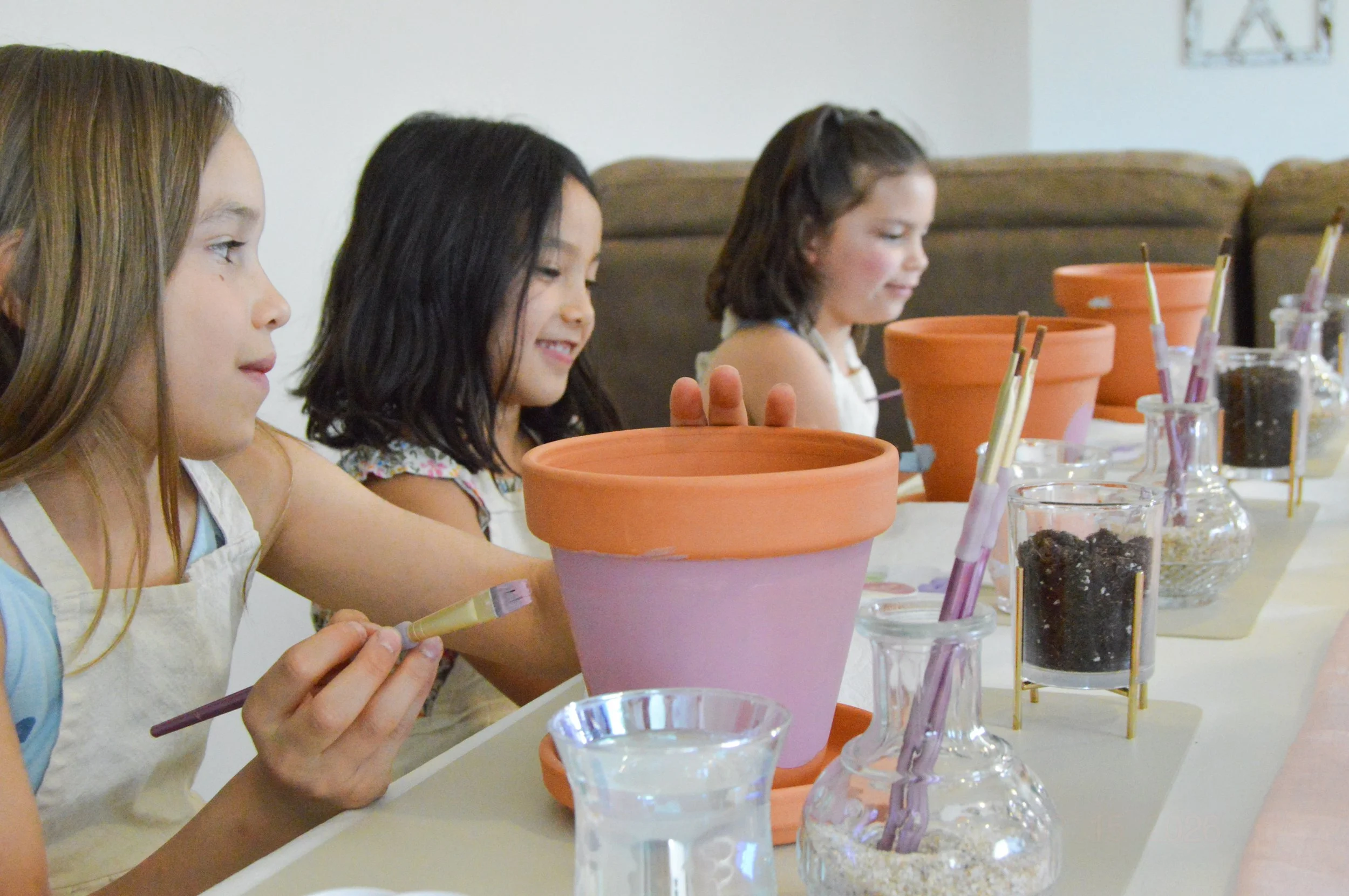 Three young girls sit at a table, each with a large clay flowerpot and various painting supplies, preparing for a pottery painting activity.
