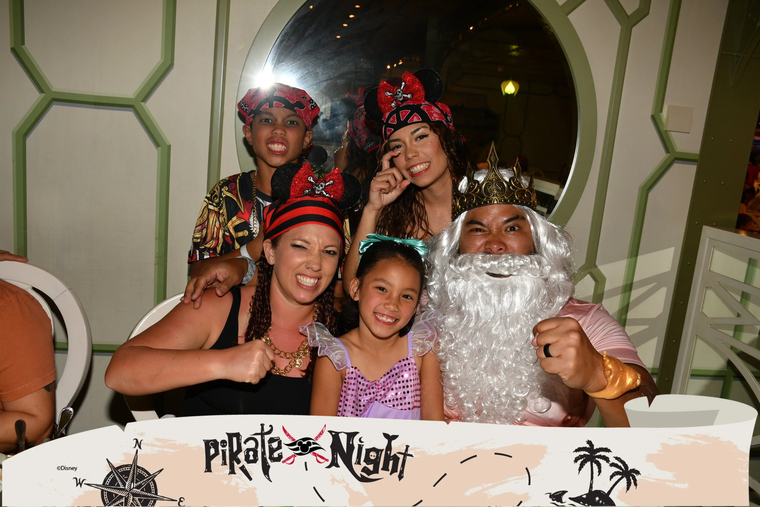 Group of people dressed as pirates and a king posing for a photo at a Pirate Night event. The group includes two women, two children, and a man dressed as a king with a crown and white beard. The background features a green wall with a mirror.