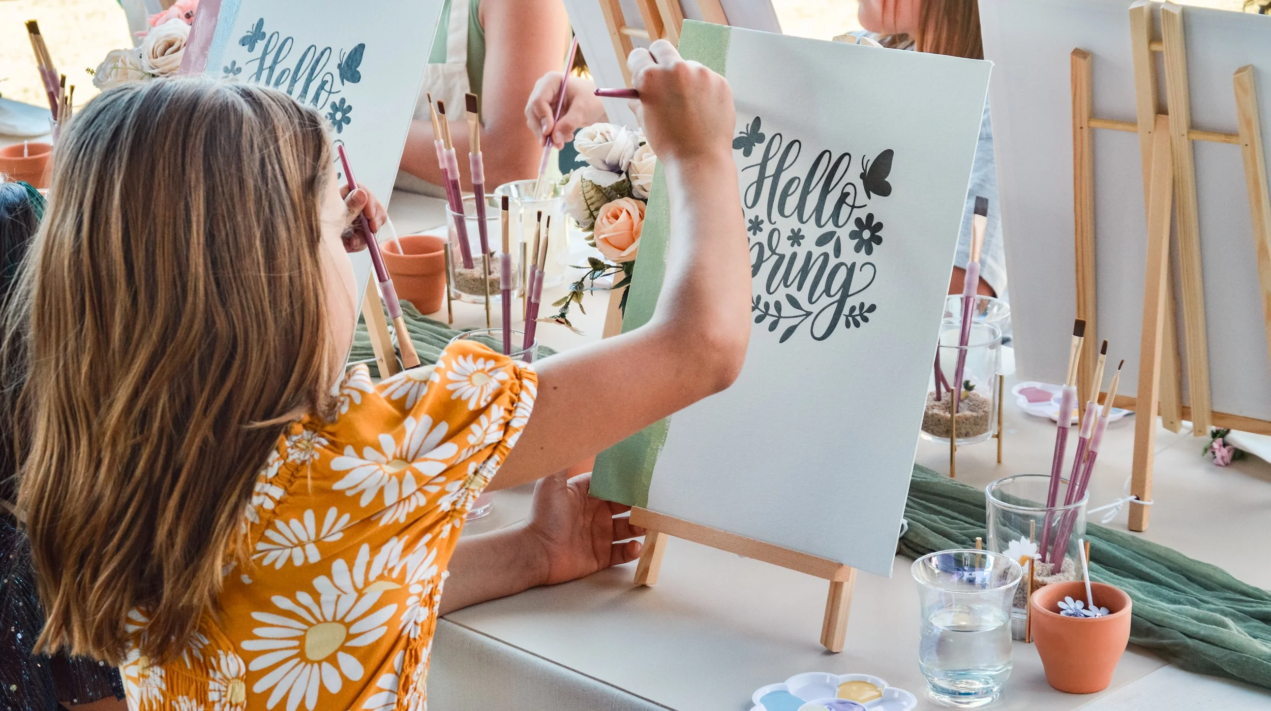 Children at a painting class, painting on canvases with 'Hello Spring' written in decorative font and floral designs.