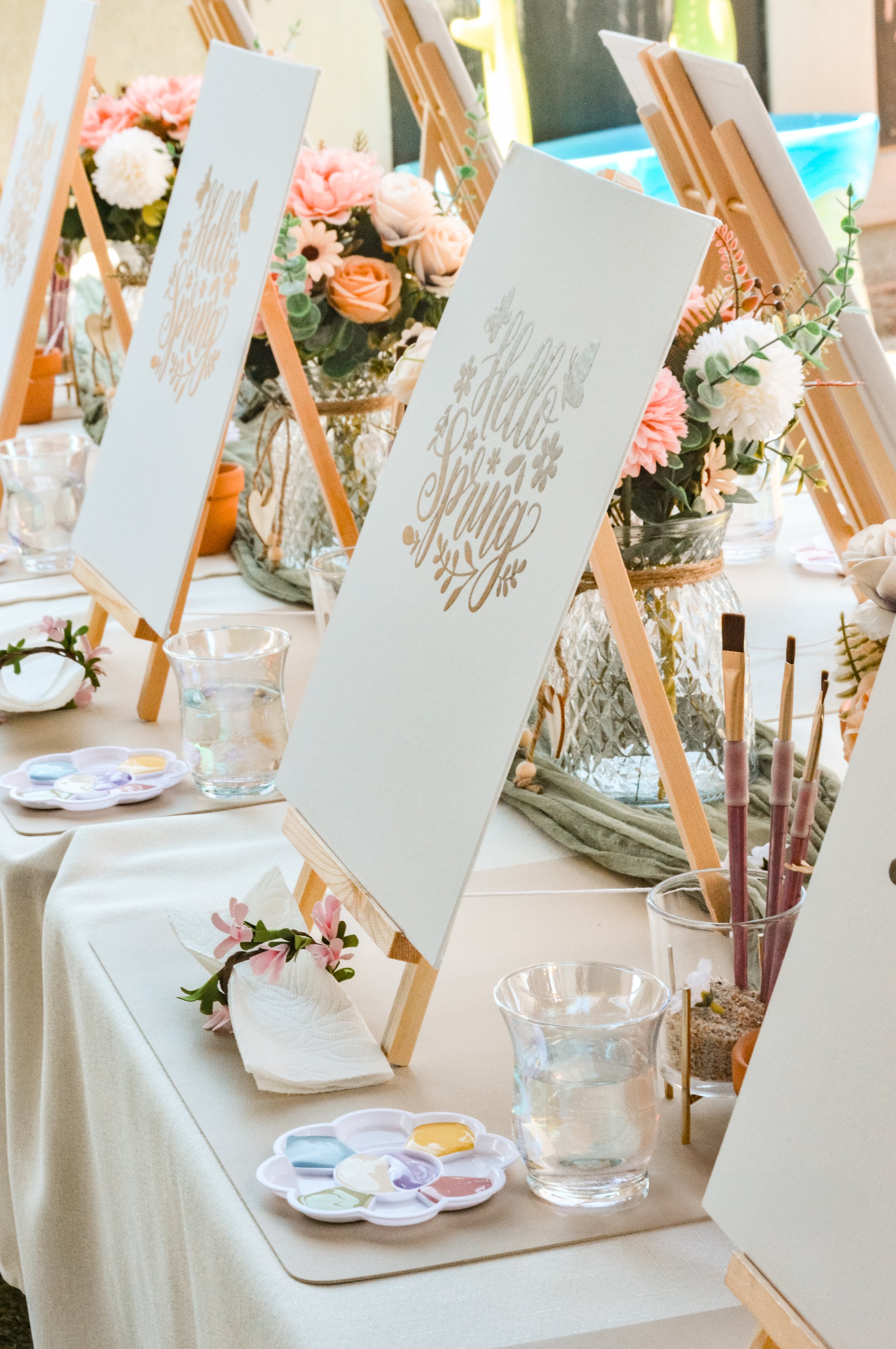 A decorated table for a spring event featuring floral arrangements, easels with signs, water glasses, paint palettes, and art supplies.