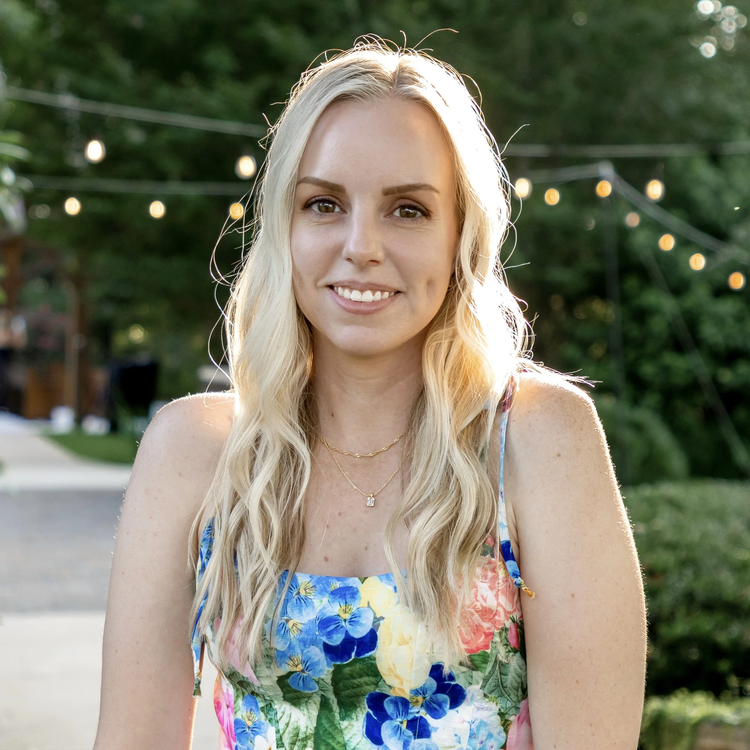 A young woman with long blonde hair, smiling, wearing a colorful floral dress, outdoors during sunset with string lights in the background.