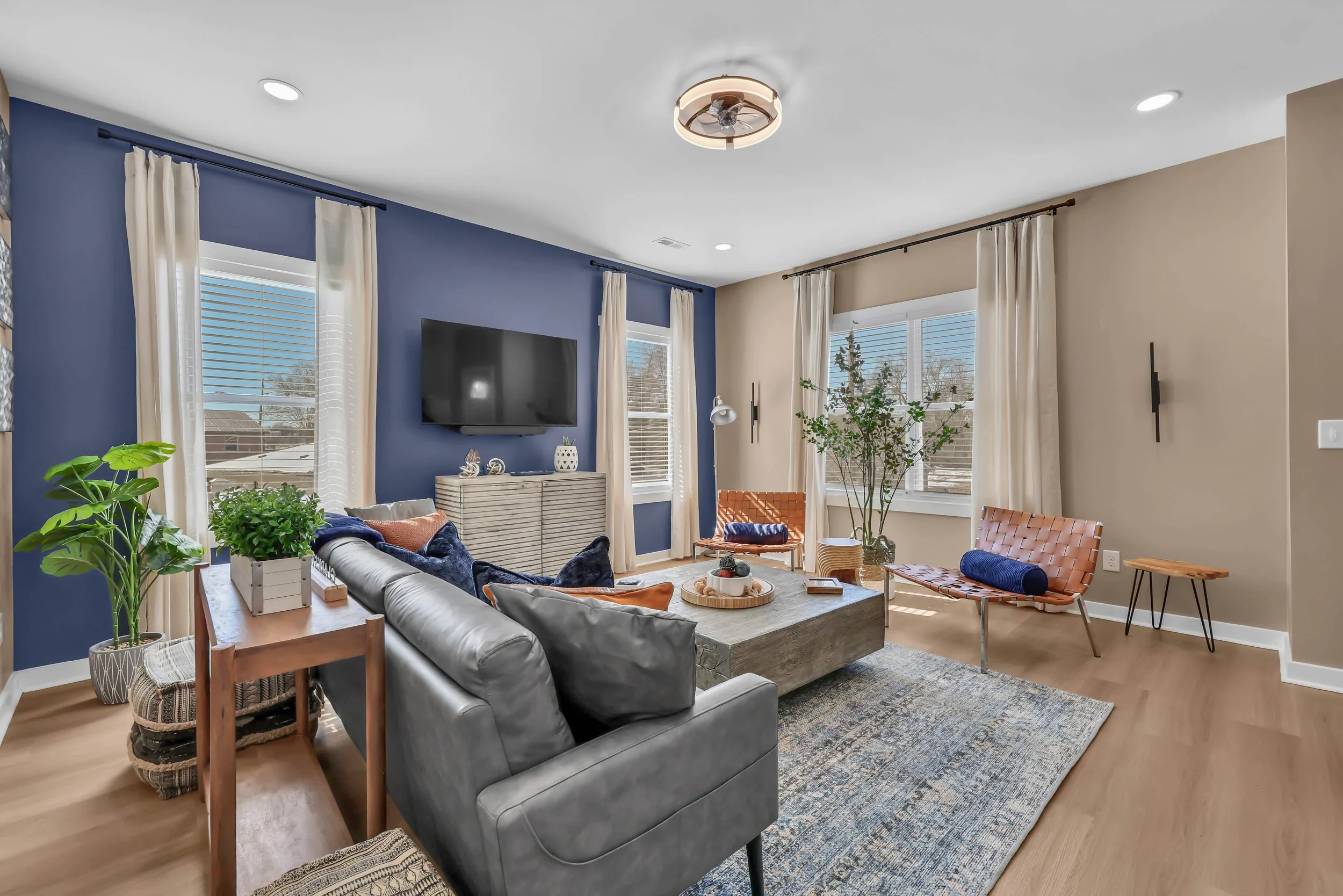 Living room with a gray leather sofa, orange and blue accent pillows, a wooden coffee table, blue and beige walls, large windows with white curtains, a wall-mounted TV, green potted plants, and modern furniture.