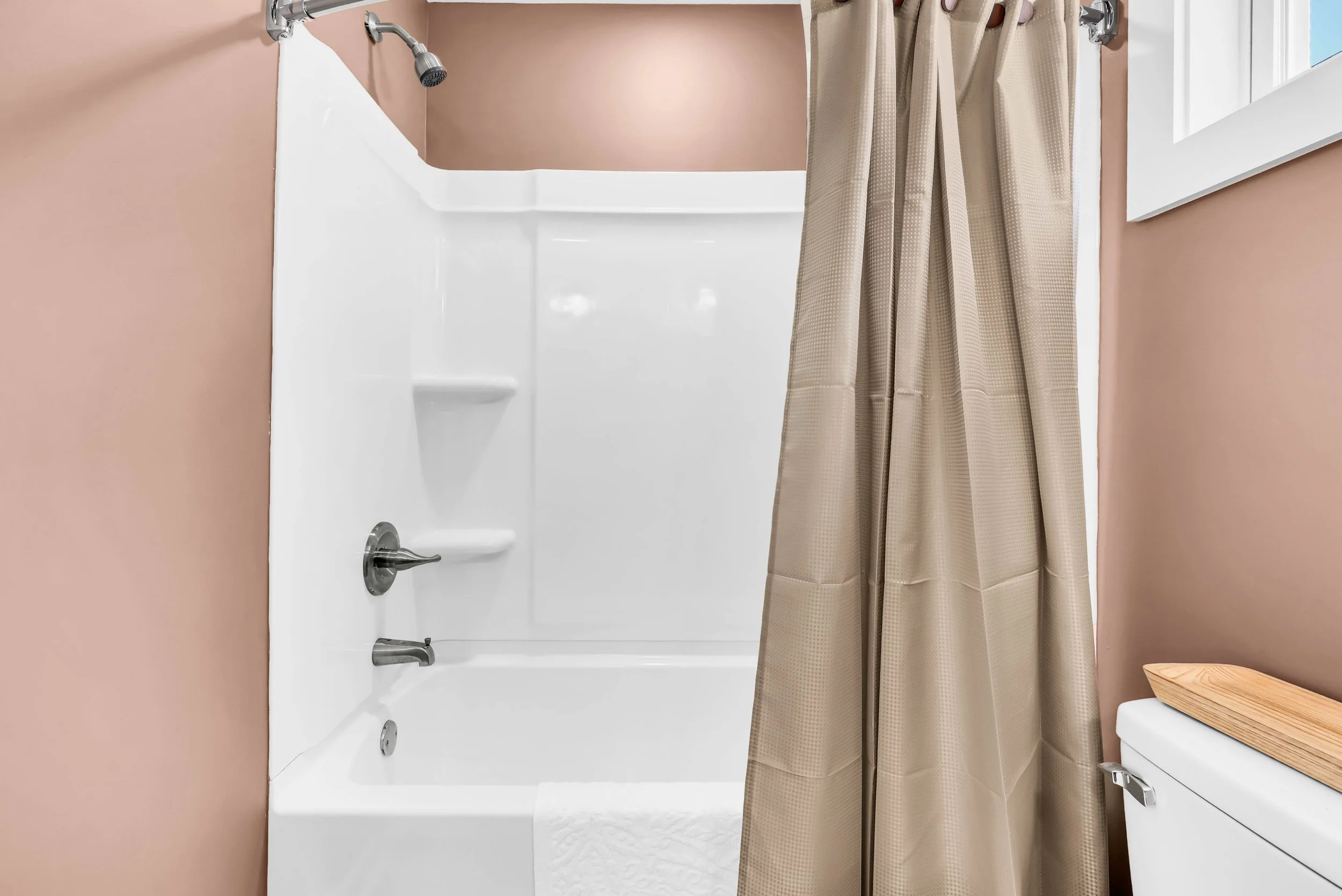 Bathroom with a bathtub and beige shower curtain, pink walls, and a window.