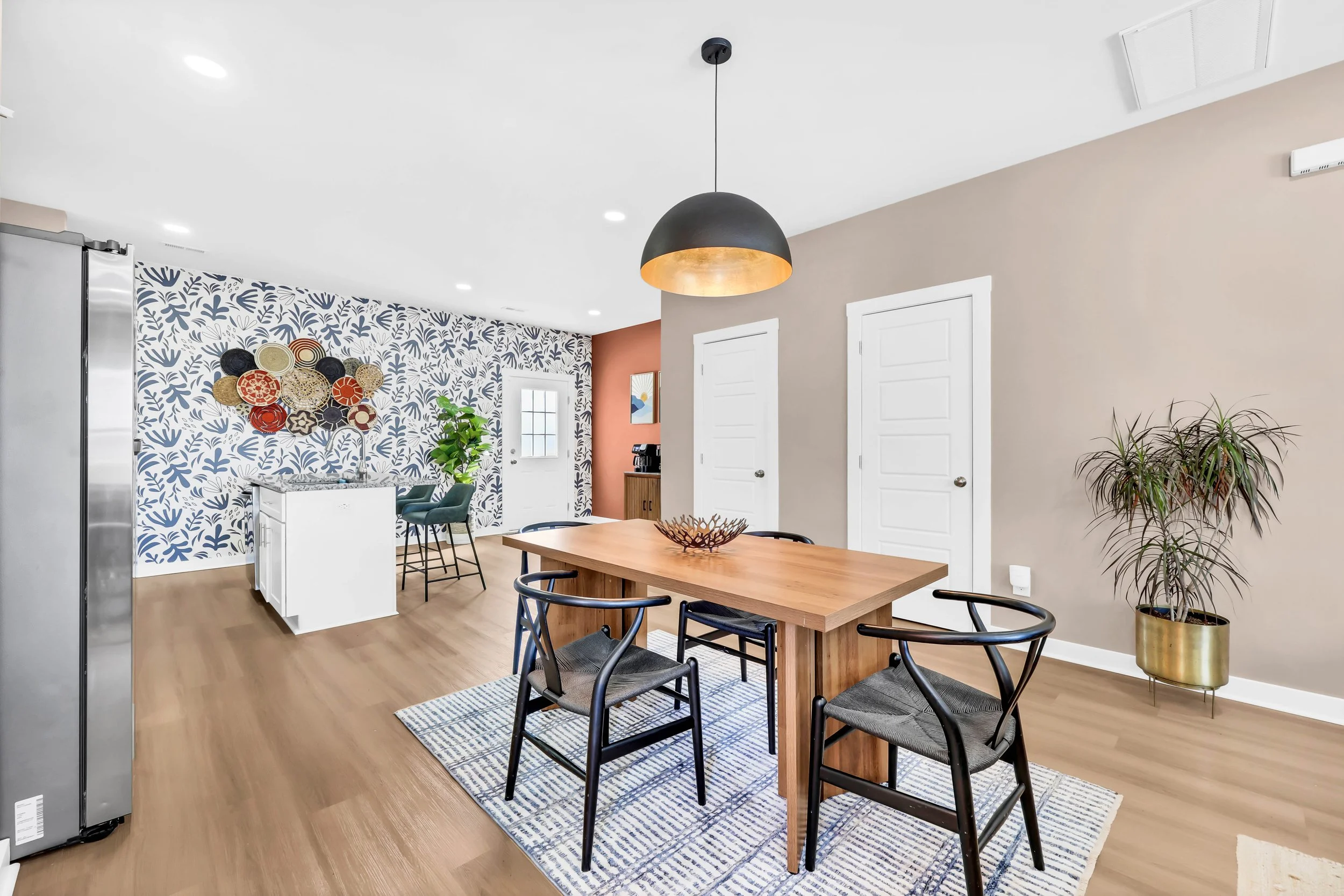 Interior of a modern dining area with a wooden table, four black chairs, a pendant light, and a patterned accent wall with wall art. There is a small kitchen island with barstools and a coffee station in the background, along with indoor plants and a hardwood floor.