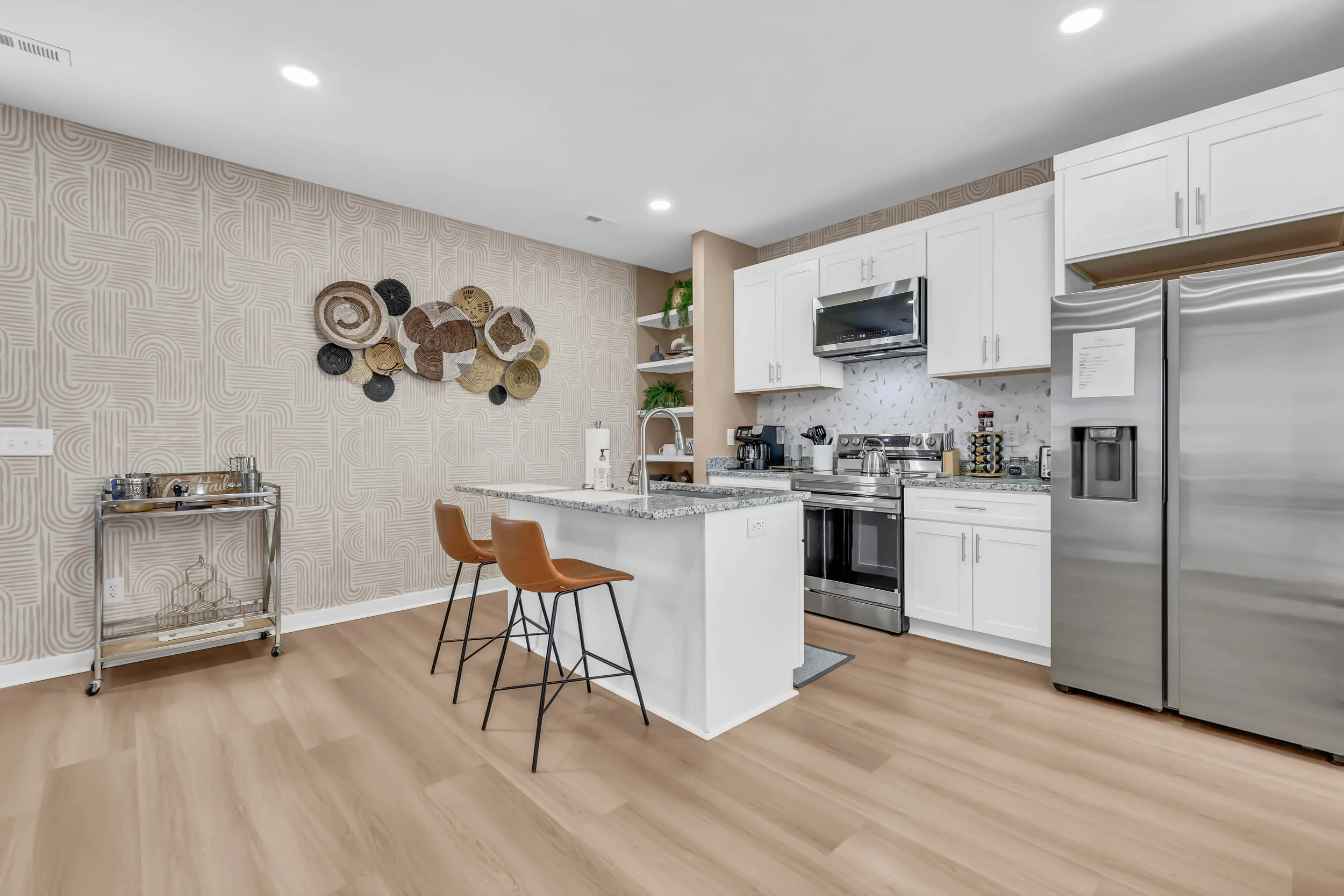 Modern kitchen with white cabinets, stainless steel appliances, granite countertop island with two brown bar stools, and decorative wall art of woven hats on a beige patterned wall.