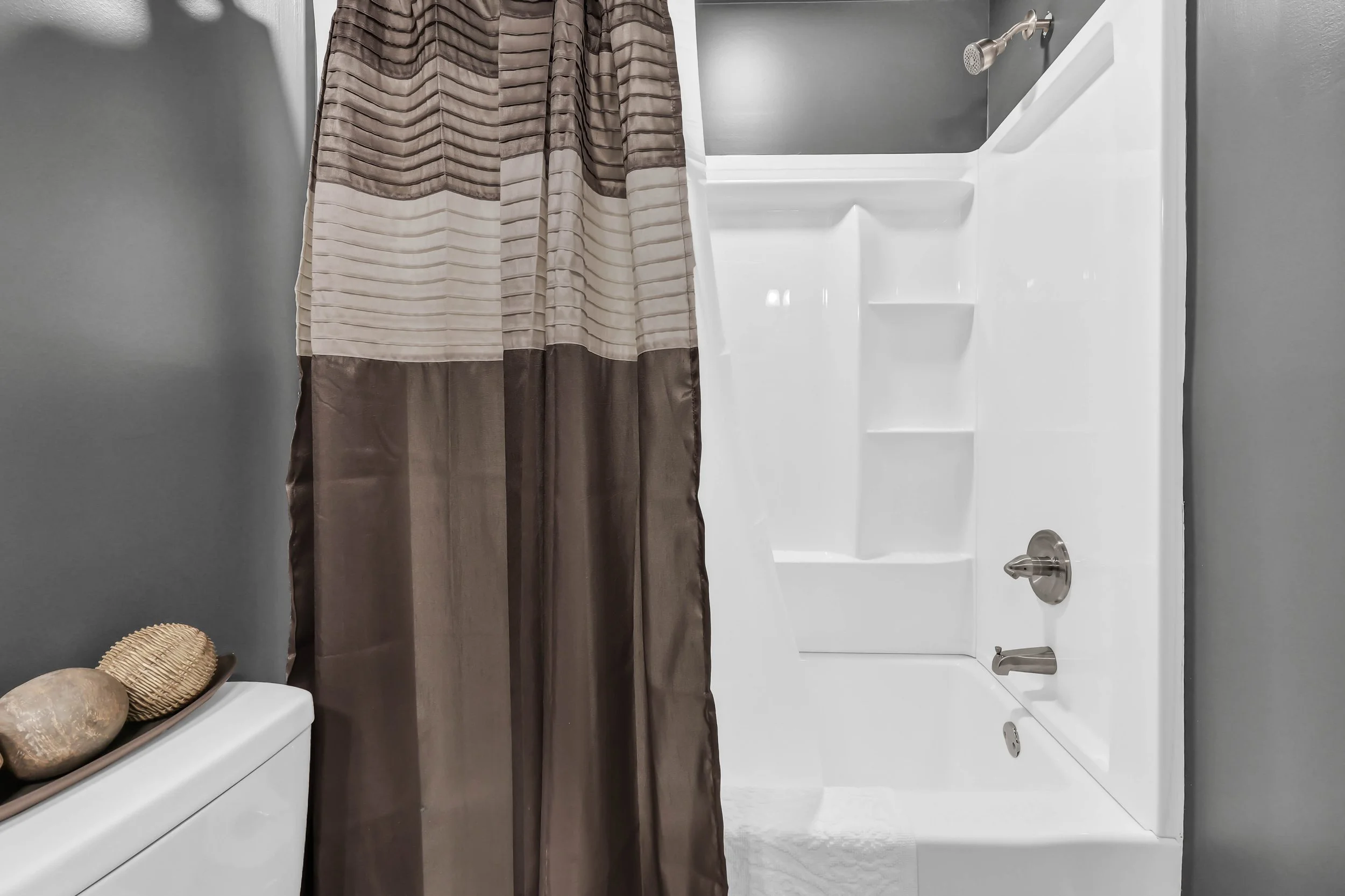 Bathroom shower with gray walls, a white shower tub assembly, a silver showerhead, and a brown and beige striped shower curtain. Part of a toilet with decorative stones on top is visible on the left.