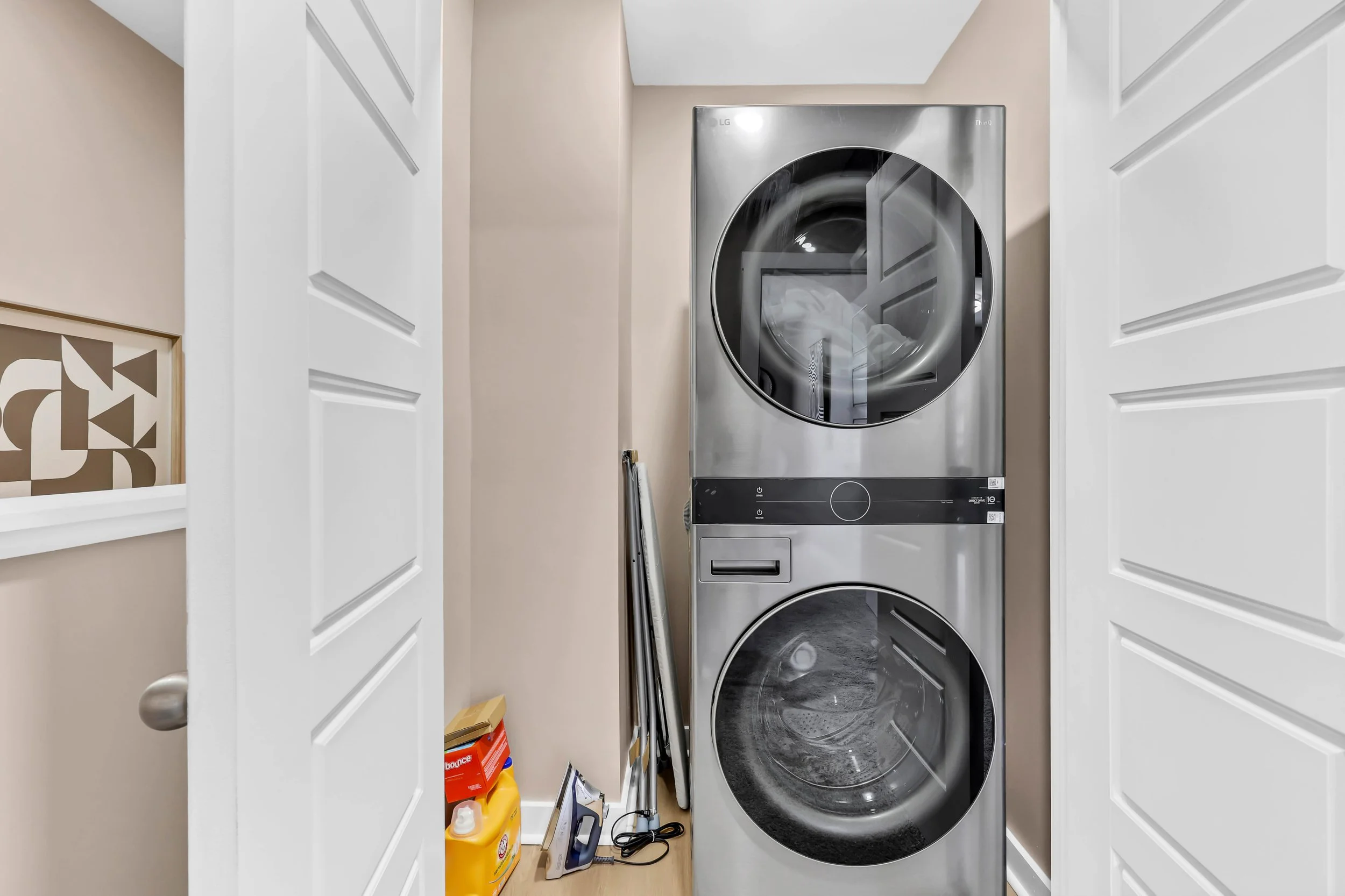 Stacked front-loading washer and dryer in a laundry room, partially closed white doors on either side, with cleaning supplies and ironing tools on the floor nearby.