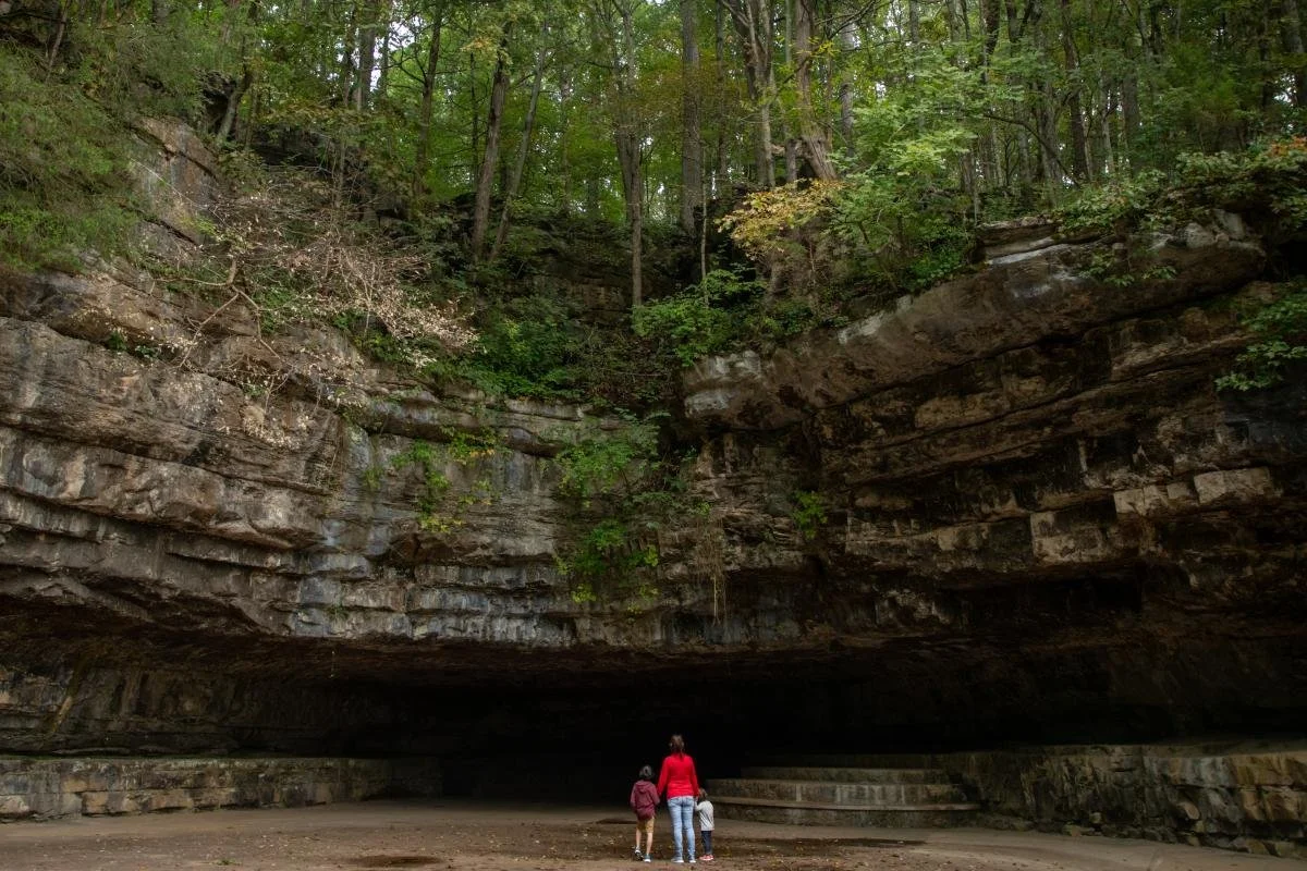 A woman and two children standing underneath a large, overhanging rock formation in a forested area with green trees overhead.