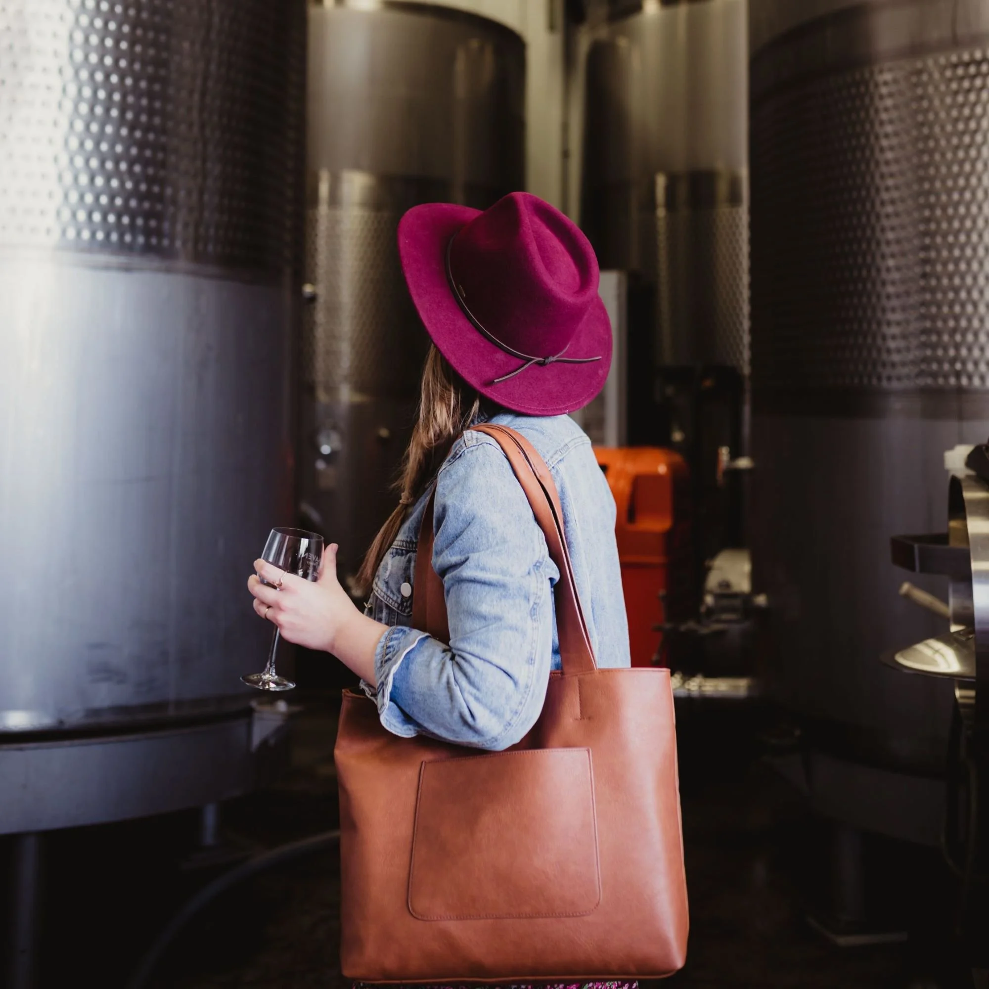 A woman with long hair wearing a burgundy hat, denim jacket, carrying a large brown leather tote bag, holding a glass of wine, standing near stainless steel wine tanks.