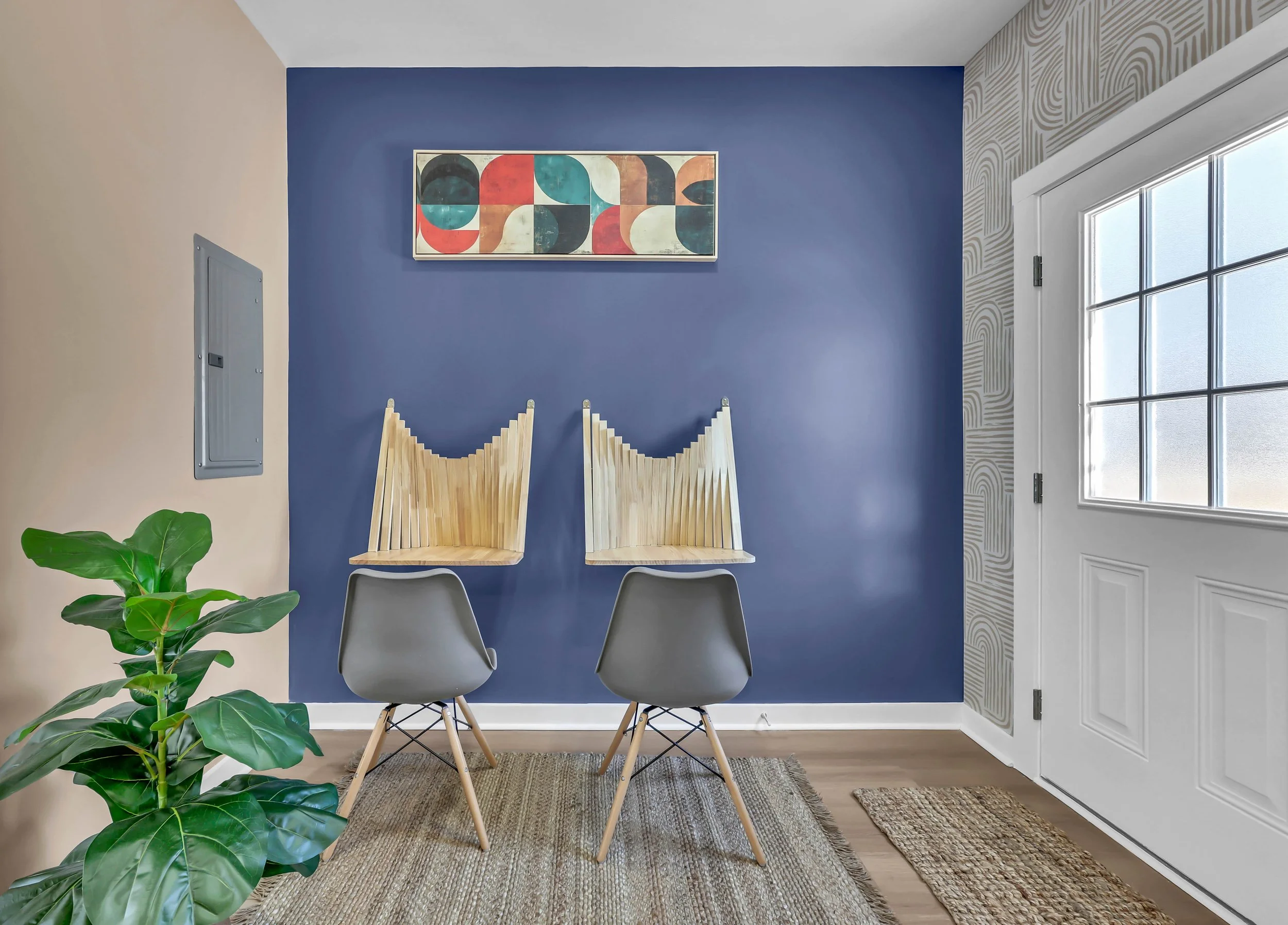 Entryway with a dark blue accent wall, two wooden wall-mounted chairs, two gray modern chairs, a patterned rug, a potted plant, a colorful abstract art piece, and a white door with window panes.