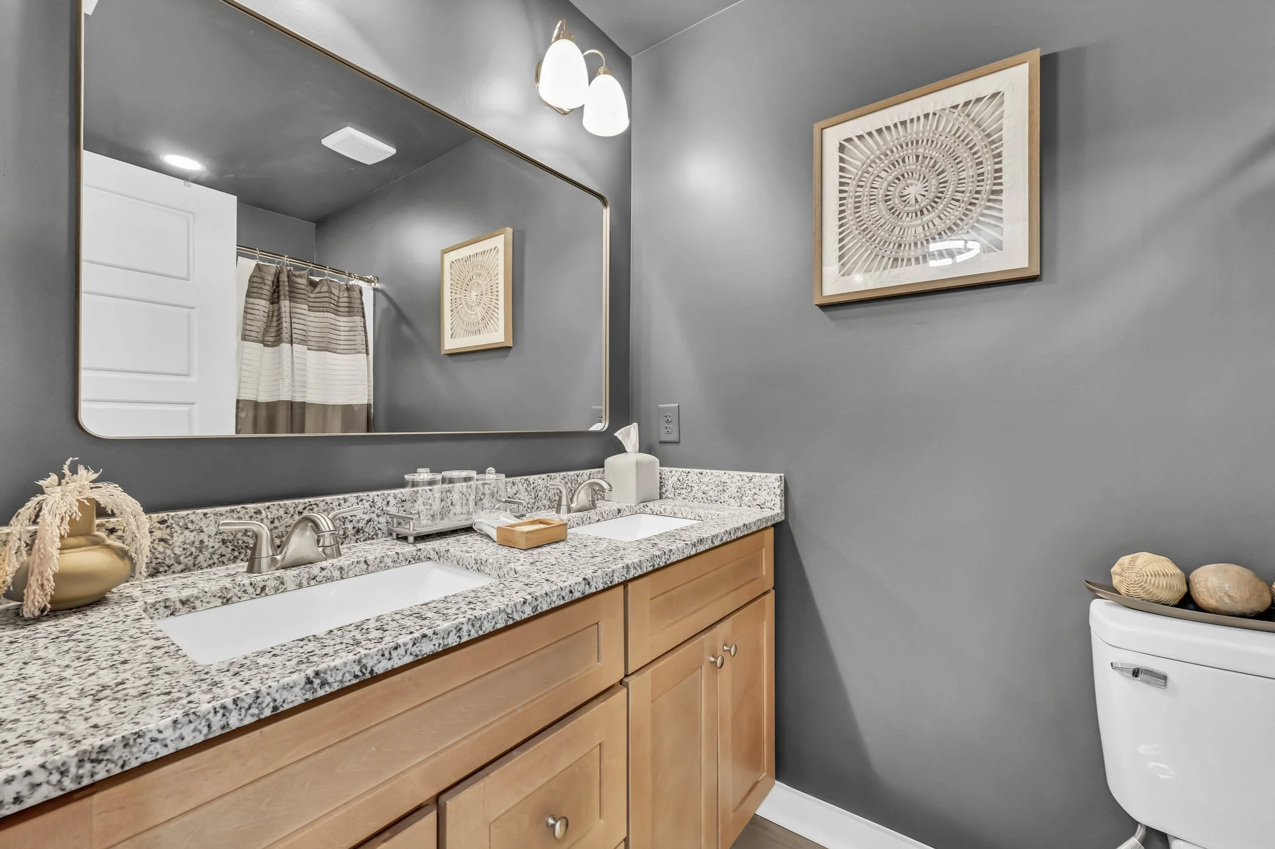 Bathroom vanity with granite countertop, double sinks, a large mirror, wooden cabinets, and a wall with gray paint. There are decorative items on the counter and wall art above the toilet.