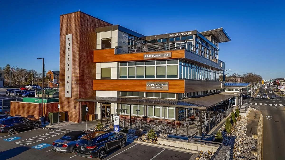 A modern multi-story commercial building with signs for various restaurants and businesses, including Skyline 500 Bar, Trattoria di Cat, and Joe's Garage, situated near a parking lot and busy street.