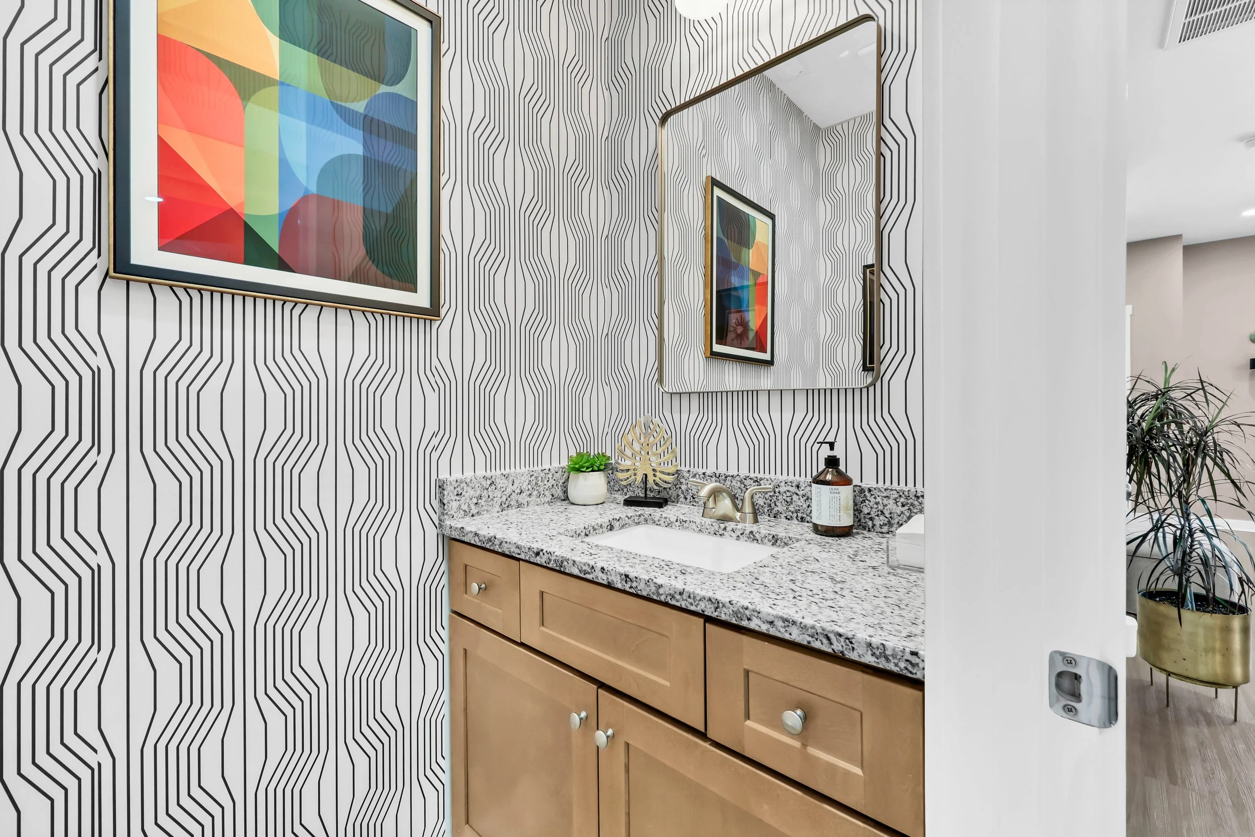 Bathroom vanity with a granite countertop, wooden cabinets, a faucet, a soap dispenser, a potted plant, and a decorative leaf. The wall above has black and white geometric patterned wallpaper and a framed abstract art piece, with a large mirror reflecting part of the room and a partial view of a potted plant in an adjacent area.