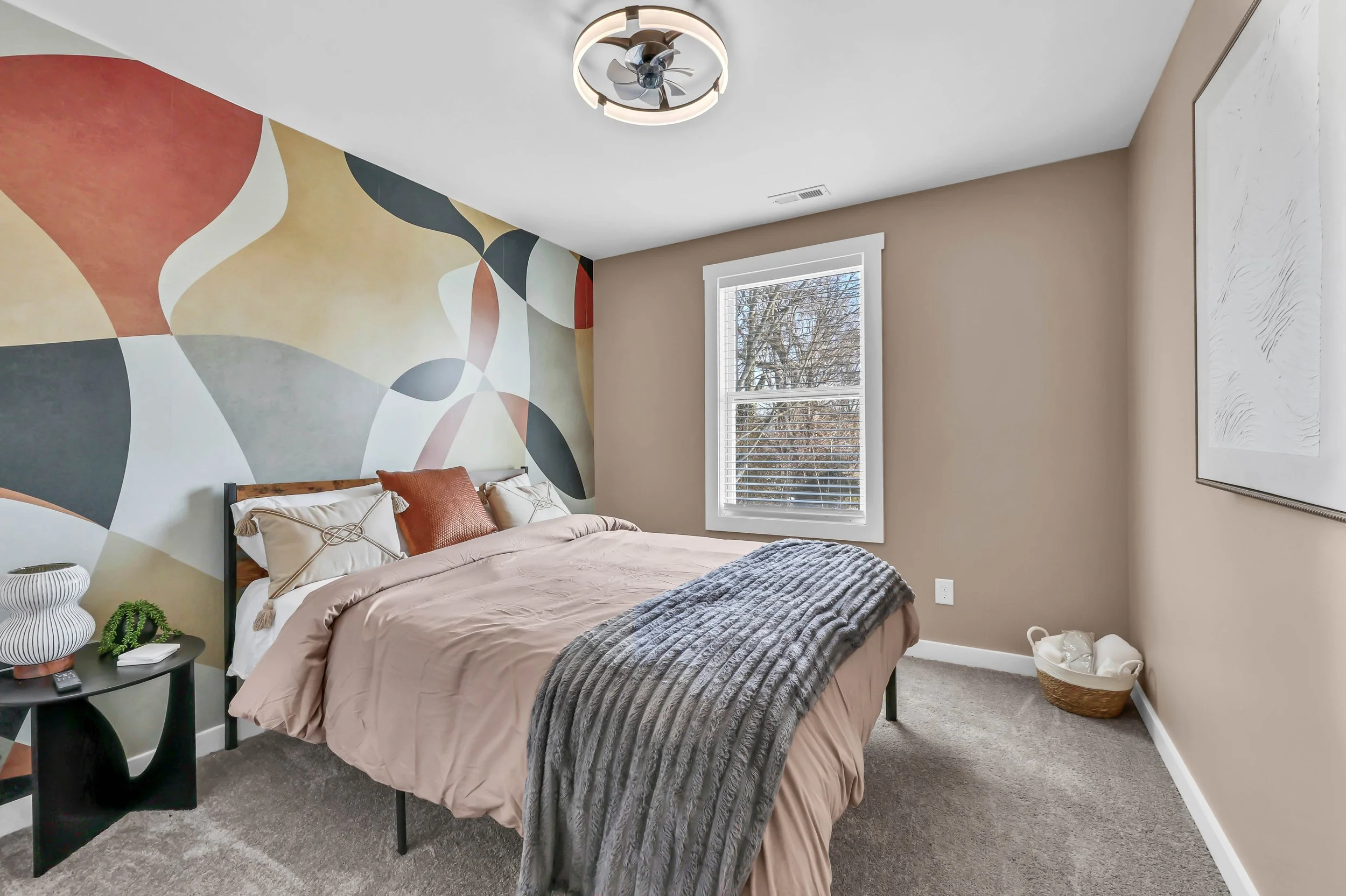 A bedroom with a bed covered in beige bedding, a gray throw blanket, and decorative pillows. There is a black nightstand with a white vase and a small plant. The room has a colorful, abstract mural on one wall, a window with white blinds, and a modern ceiling fan with a light fixture. A large framed artwork hangs on the wall, and a basket with rolled towels is on the floor.
