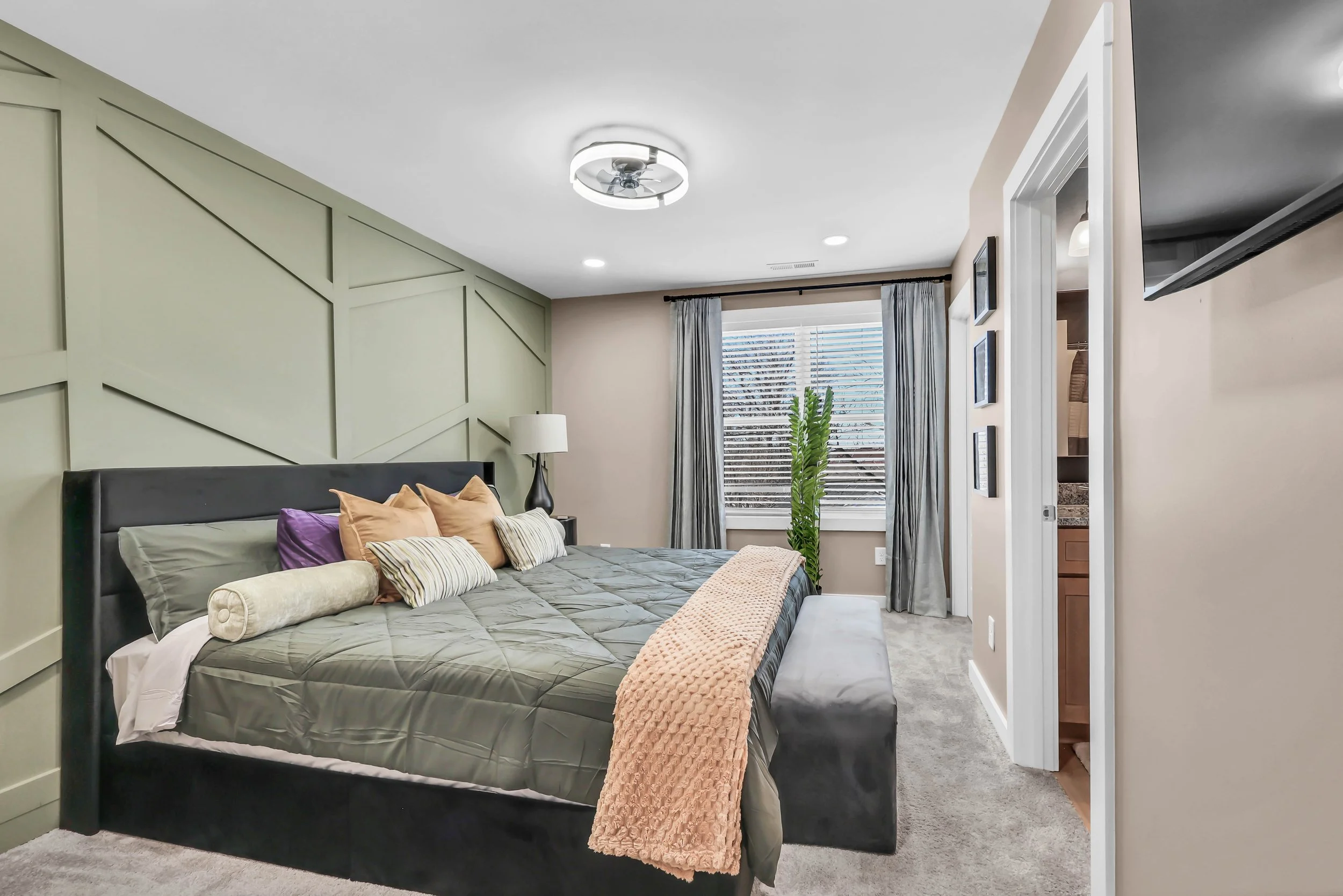 Modern bedroom with bed, decorative pillows, green accent wall, window with blinds, potted plant, and wall-mounted TV.