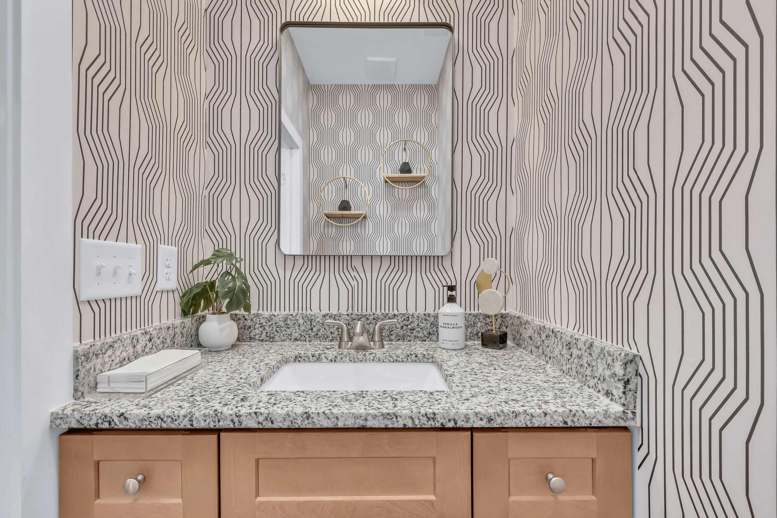 Bathroom vanity with a granite countertop, a white sink, and a mirror with decorative shelving on a geometric patterned wallpaper.