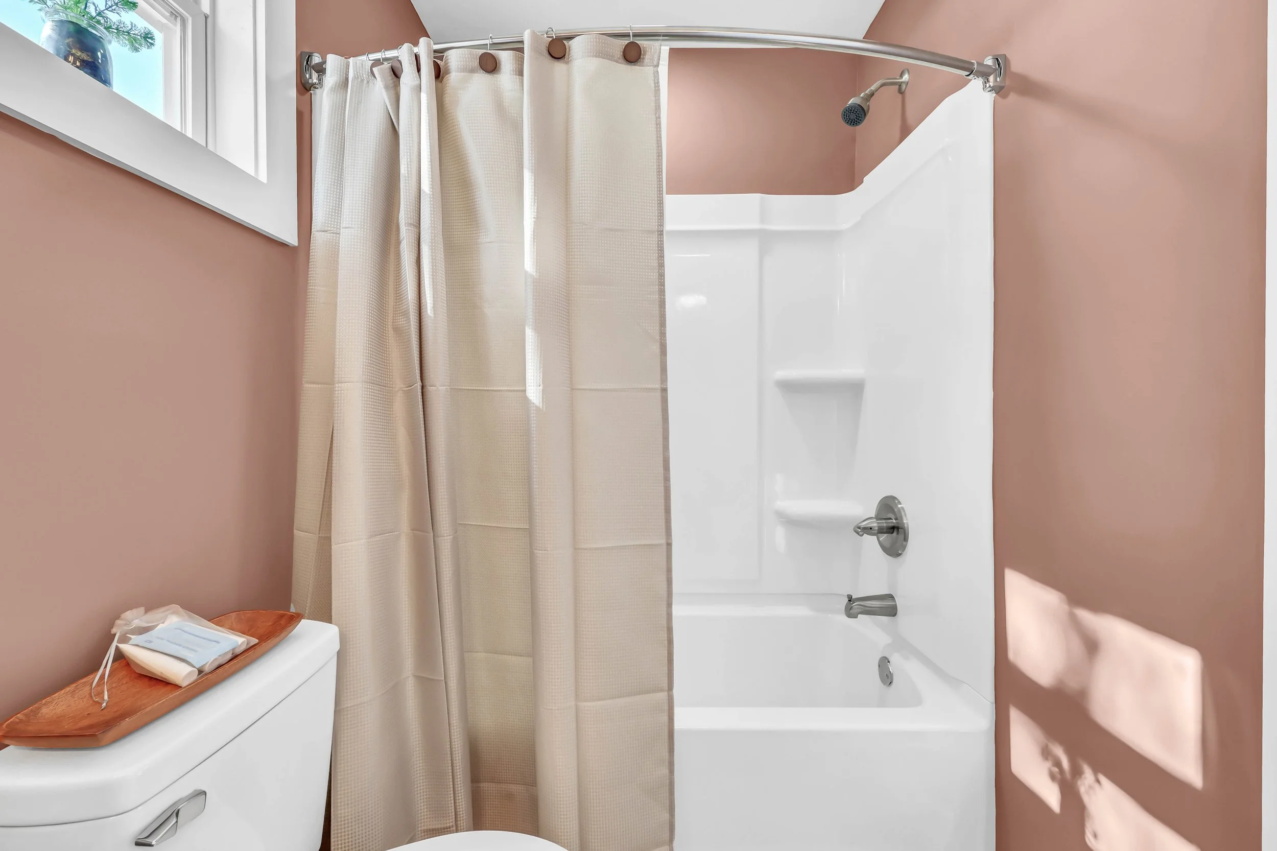 Bathroom with a shower and bathtub, tan walls, a window with a plant, a toilet with a wooden tray holding toiletries, and a cream-colored shower curtain.