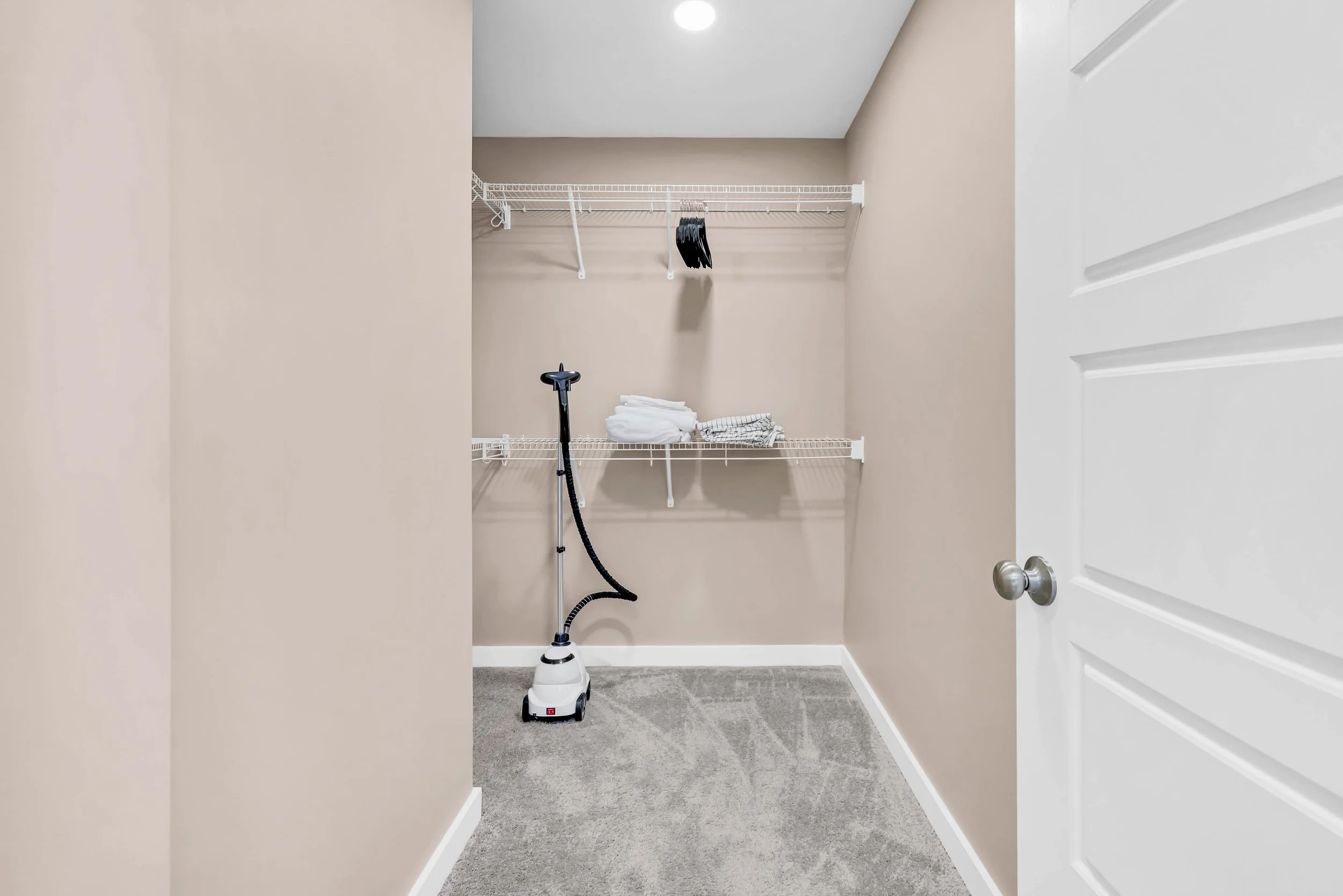 Empty walk-in closet with beige walls, gray carpet, white wire shelves, folded towels, a black hairbrush, and a handheld vacuum