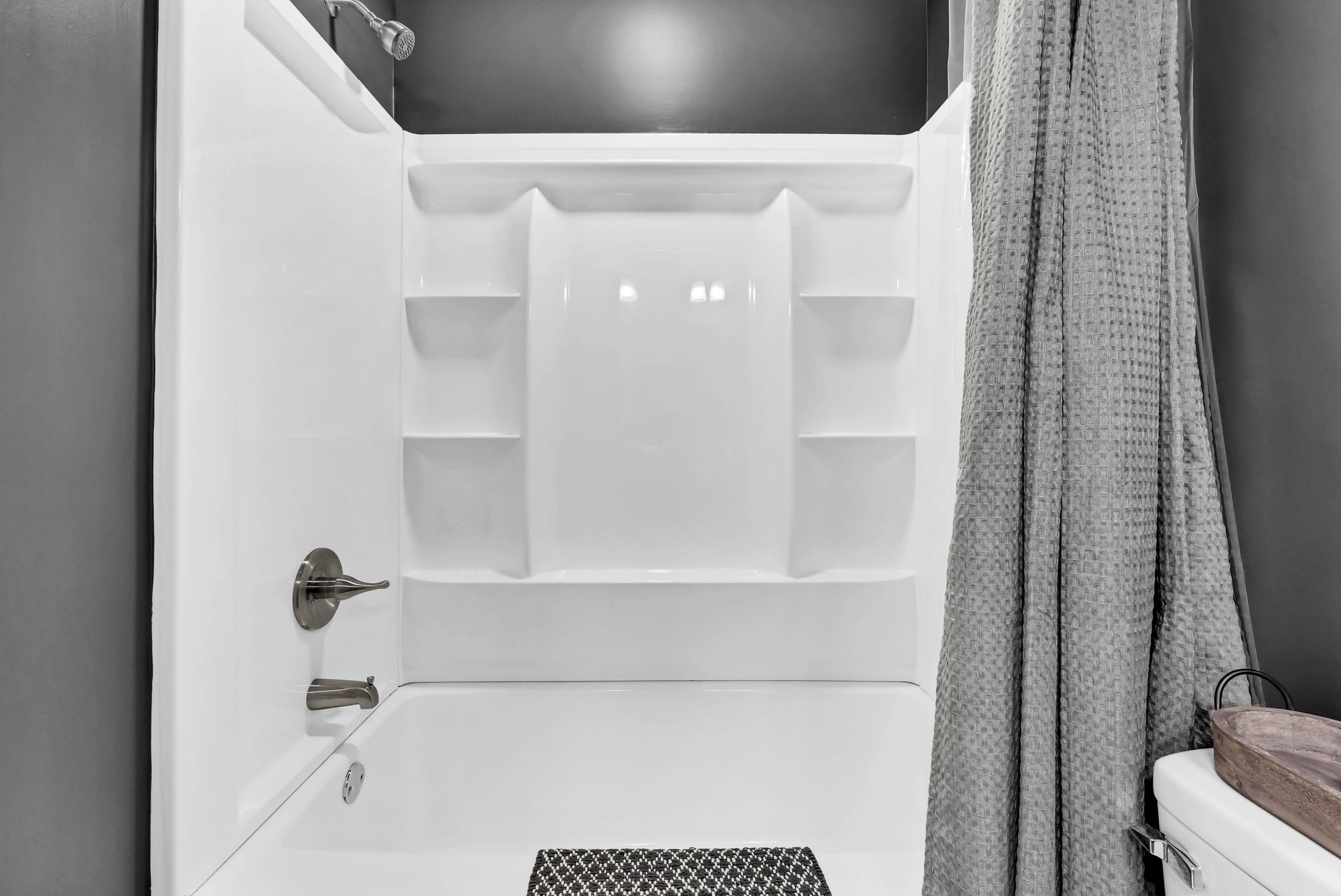 White bathtub with built-in shelves, silver faucet and showerhead, gray walls, gray textured shower curtain, and a small brown tray on top of the toilet.