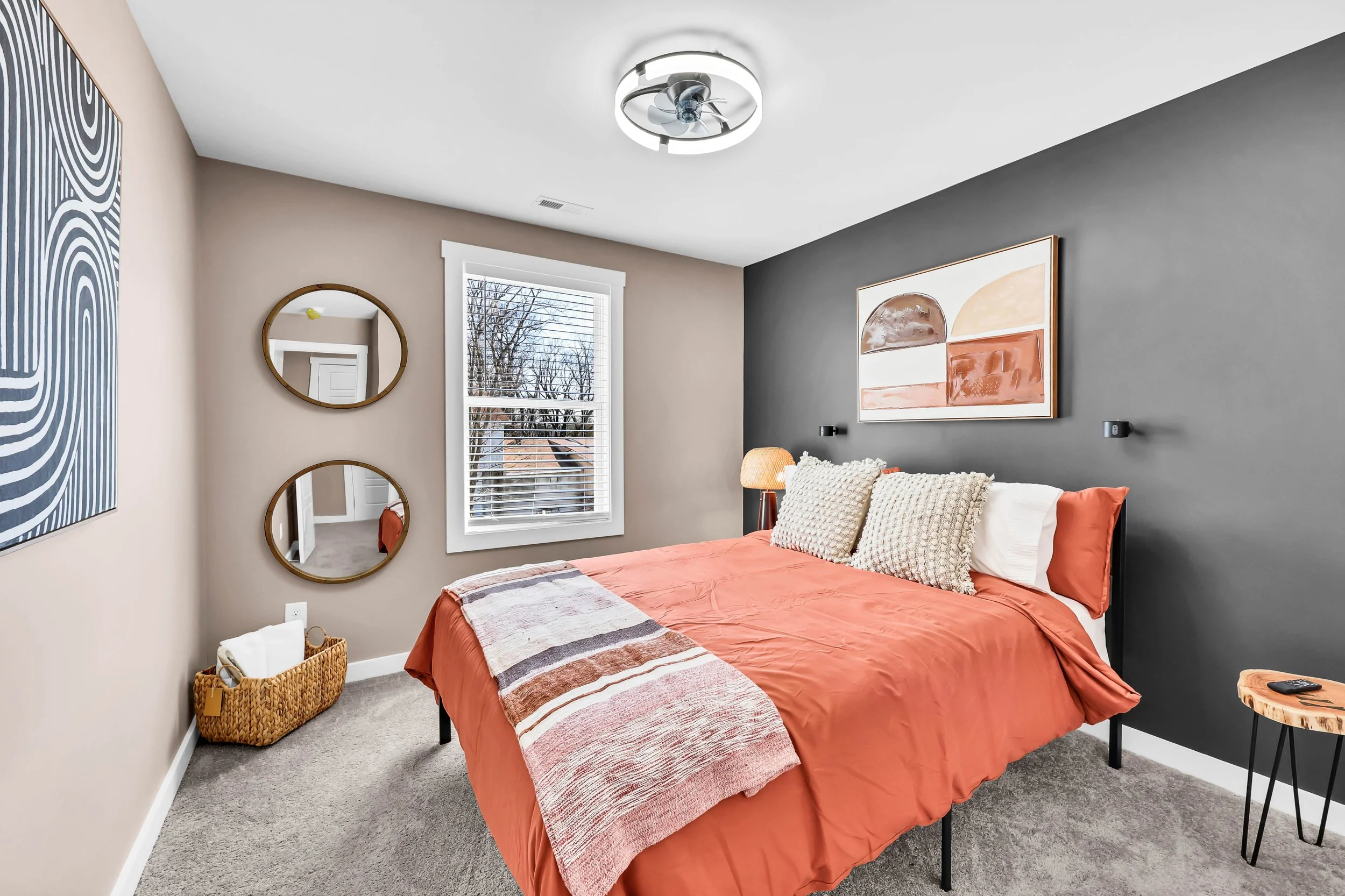 Bedroom with a bed, orange bedding, beige and white pillows, dark gray accent wall, window with blinds, artwork on walls, and a bedside table with a remote control.