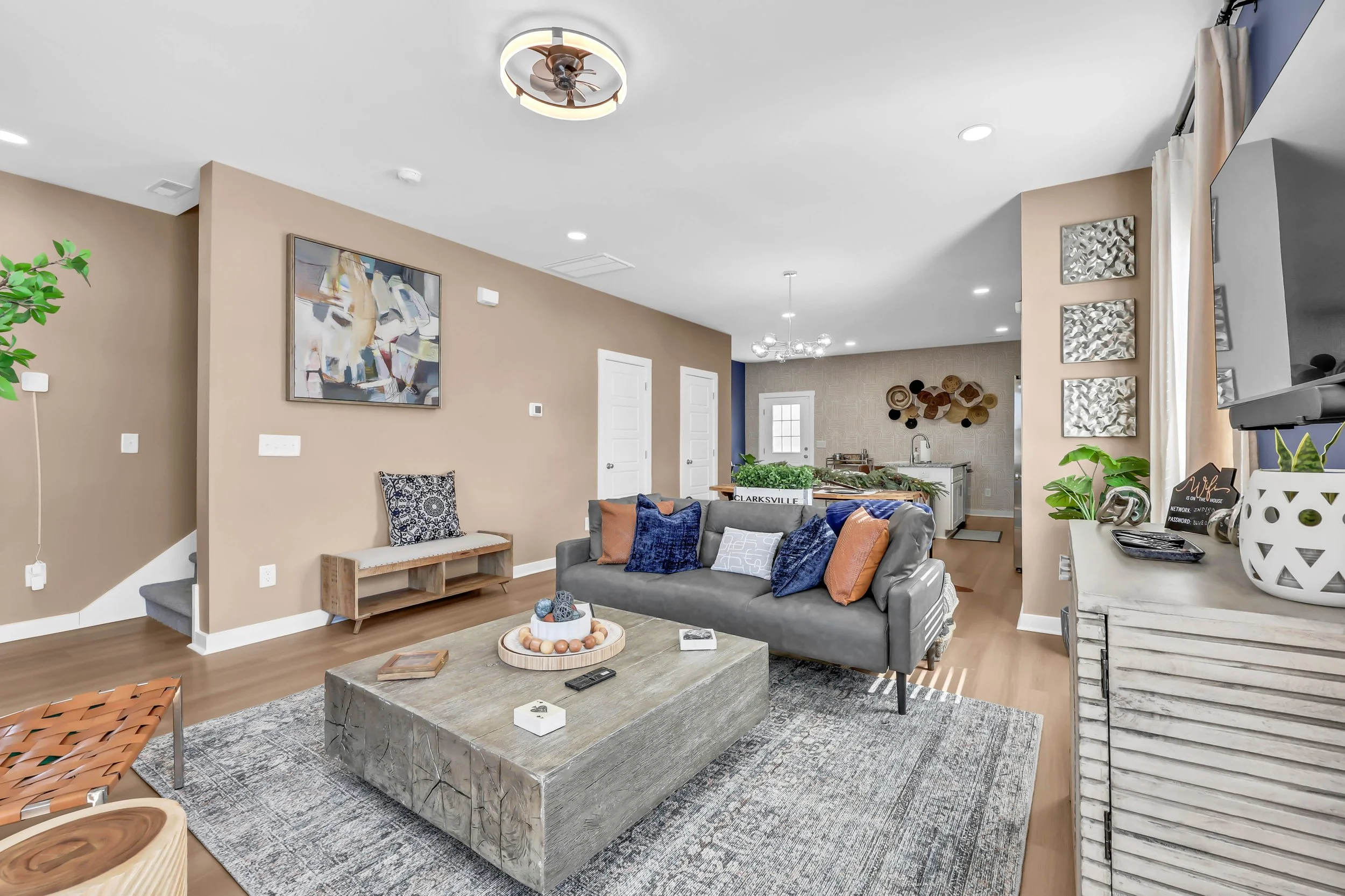 Living room with gray sofa, wooden coffee table, beige walls, colorful pillows, potted plants, wall art, and kitchen in the background.