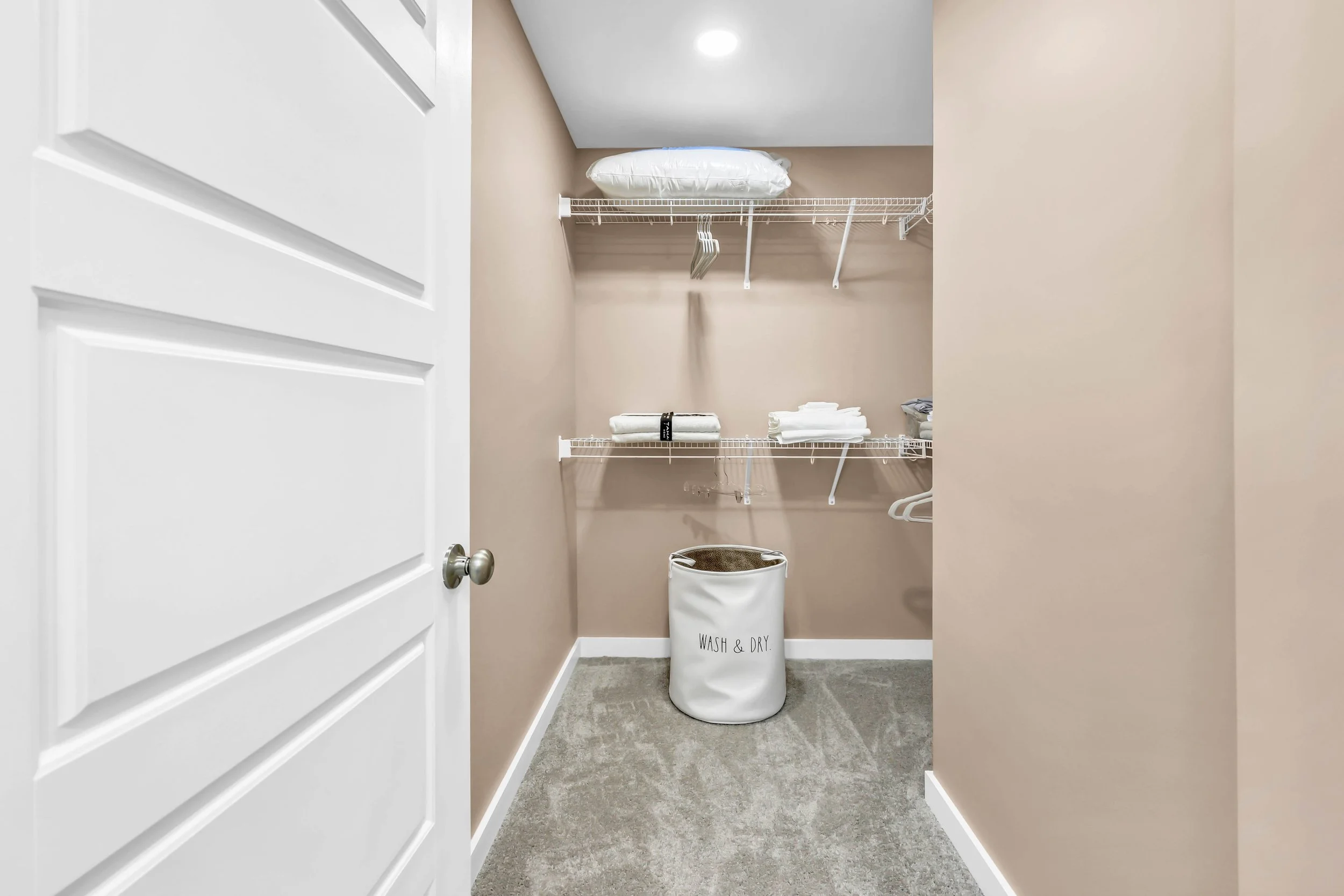 Empty closet with white wire shelves, folded towels, and a laundry basket labeled 'Wash & Dry' on a beige wall and carpeted floor.