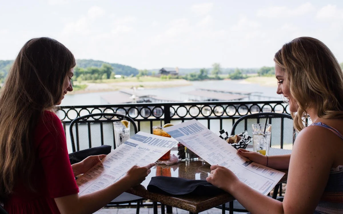 Two women sitting at an outdoor restaurant table with drinks, reading menus, with a river and boats in the background.