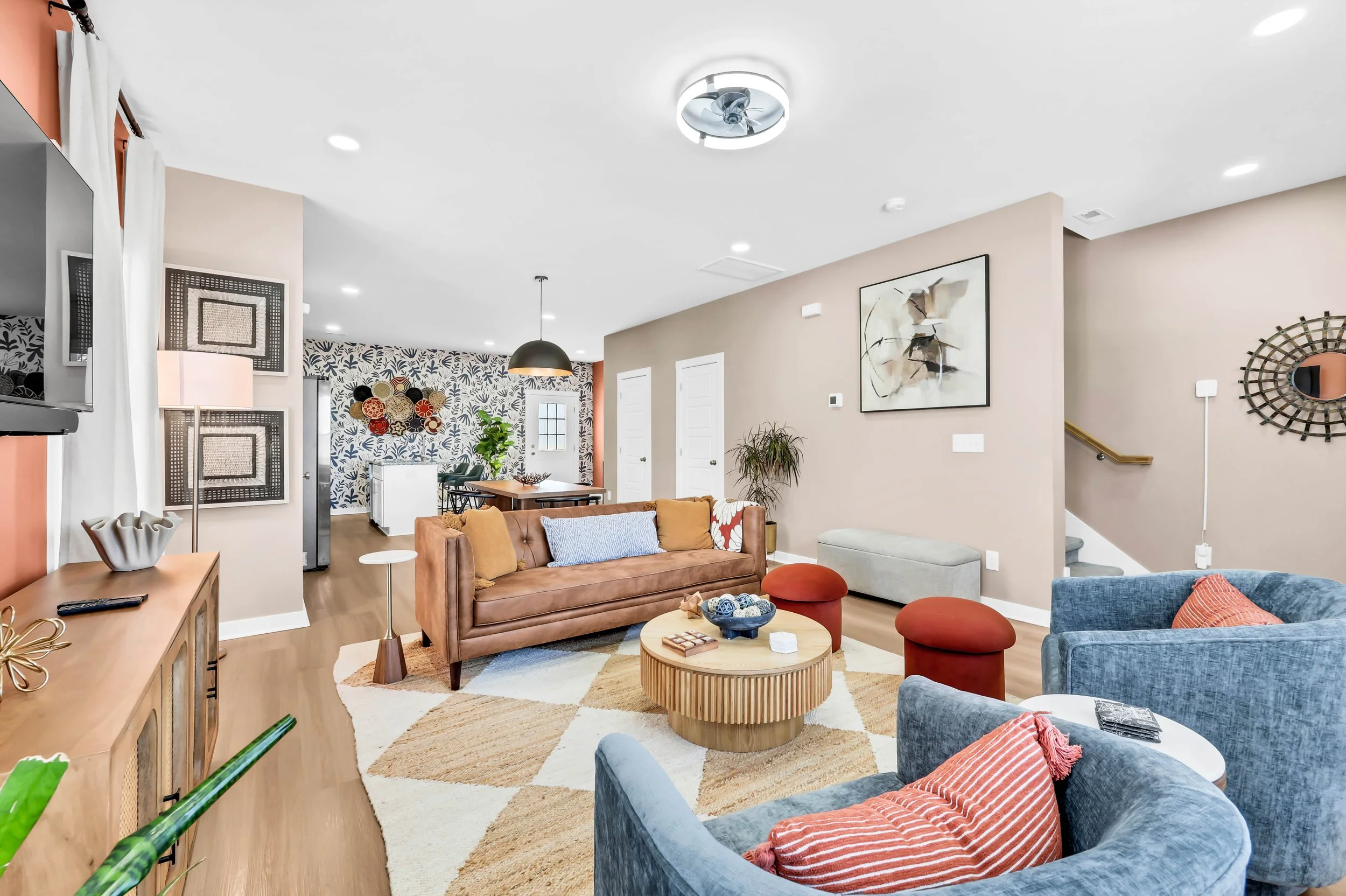 A spacious living room with beige walls, modern furniture, a tan sofa, blue armchair, orange pillows, a round wooden coffee table, and decorative items. There is a dining area in the background with a black pendant light, patterned wallpaper, and a wall hanging.