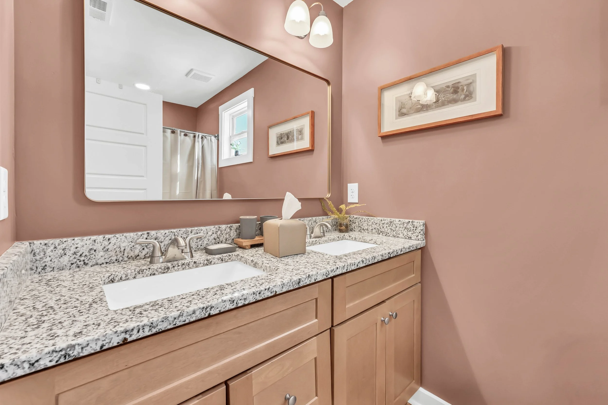 Bathroom with a double granite vanity, large mirror, framed artwork on pink walls, small window, tissue box, soap dispenser, and a decorative plant.