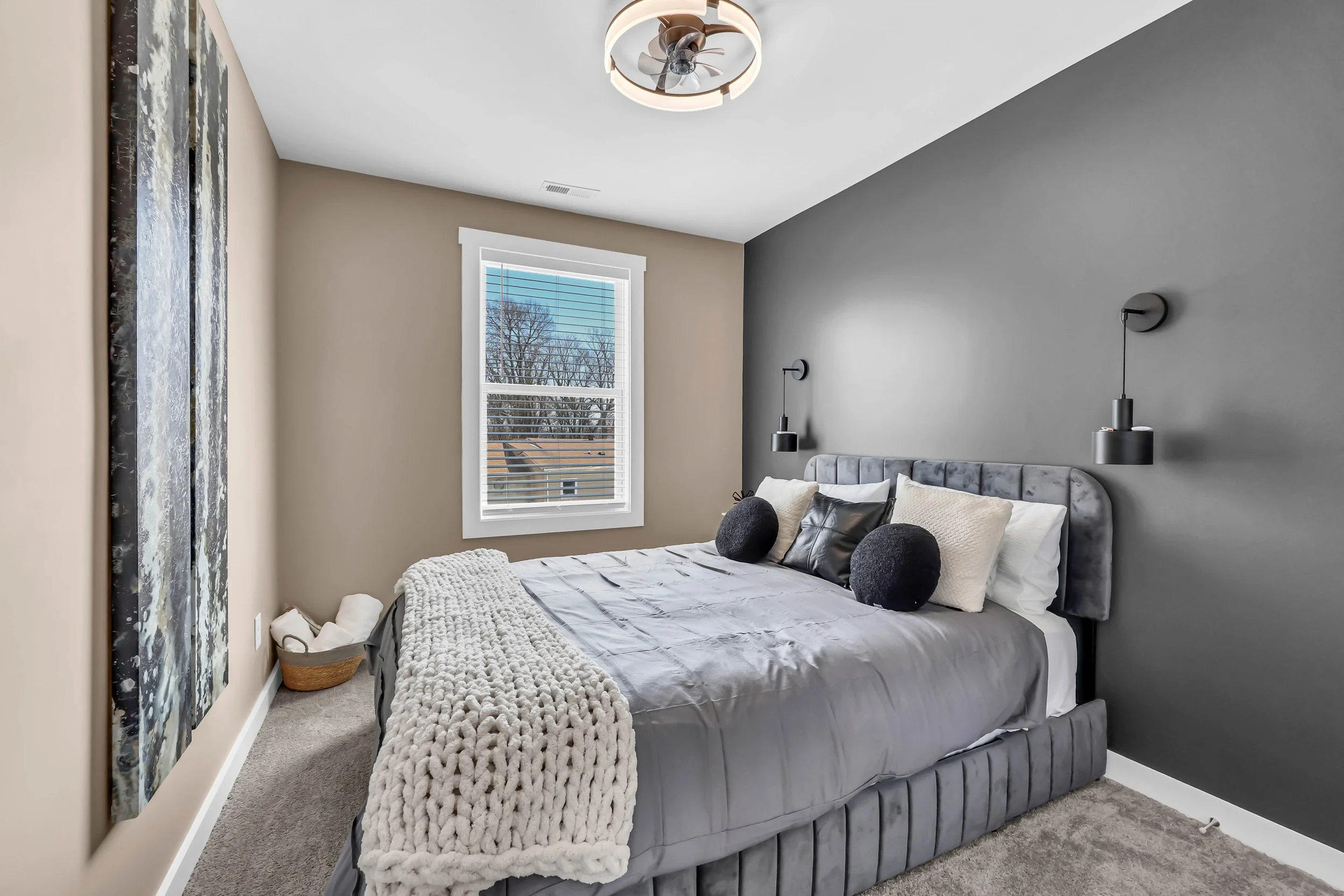 A modern bedroom with a gray upholstered bed, black and white pillows, and a chunky white blanket. Dark gray wall with two black wall-mounted reading lights, a gray ceiling fan, a window with white trim showing trees outside, and beige walls.