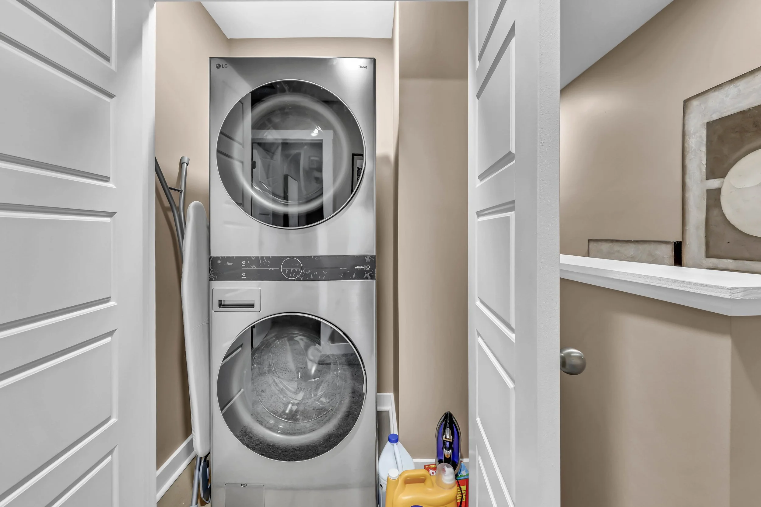 A laundry room with stacked front-loading washer and dryer, beige walls, and cleaning supplies on the floor.