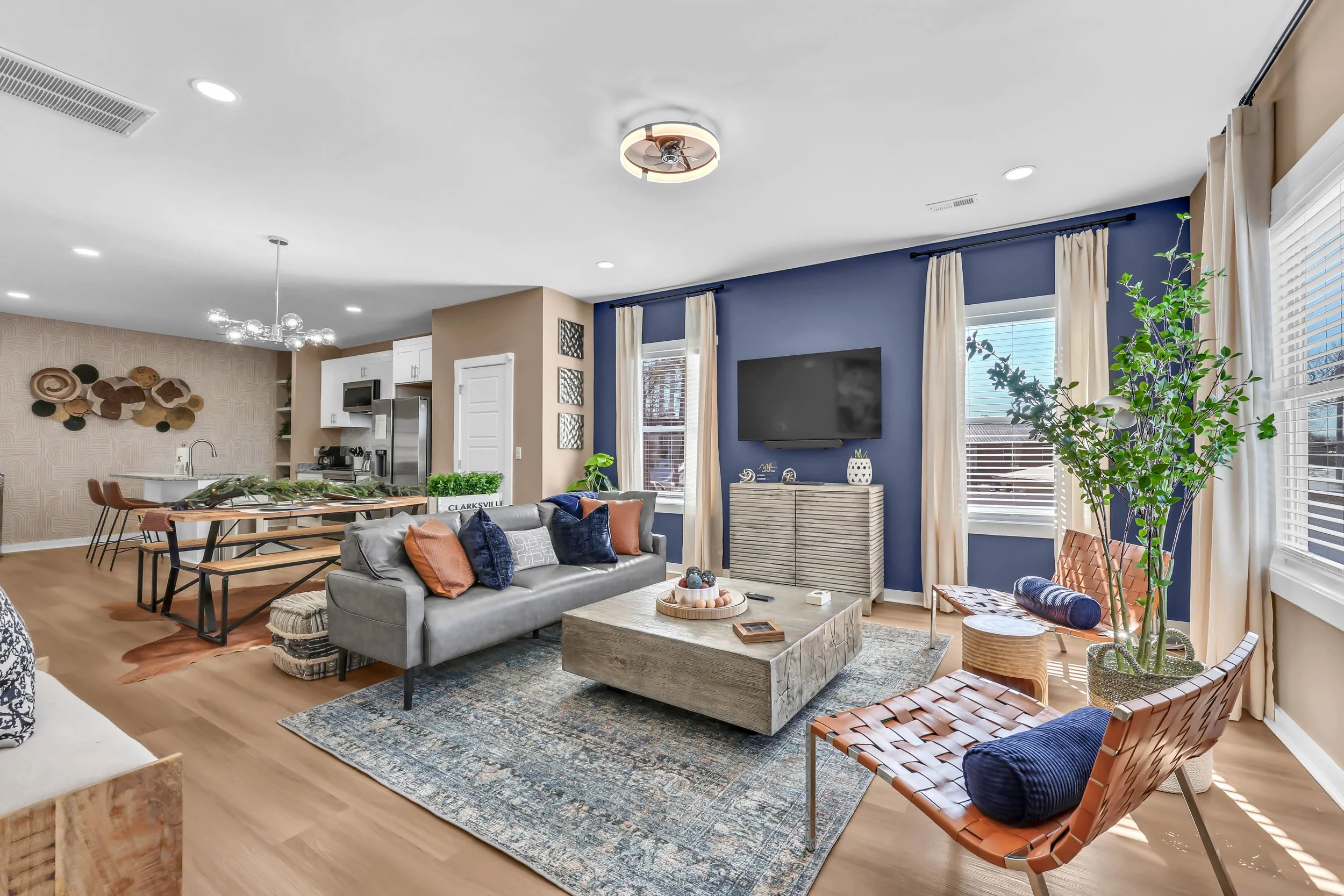 A modern living room with a blue accent wall, a mounted TV, beige curtains, a gray sofa with cushions, a wooden coffee table, leather chairs with cushions, a large patterned area rug, and a large potted plant near windows. The open kitchen and dining area are visible in the background.