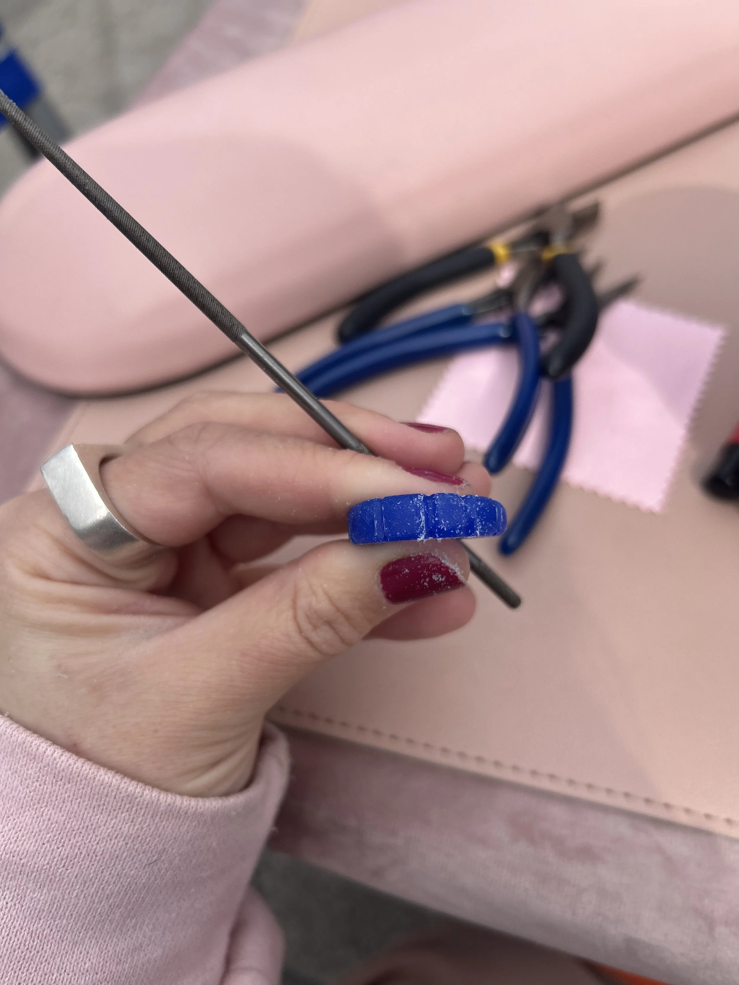 A person holding a small blue polishing wheel and a thin metal file or bur, with jewelry pliers and a pink cloth in the background, on a pink surface.