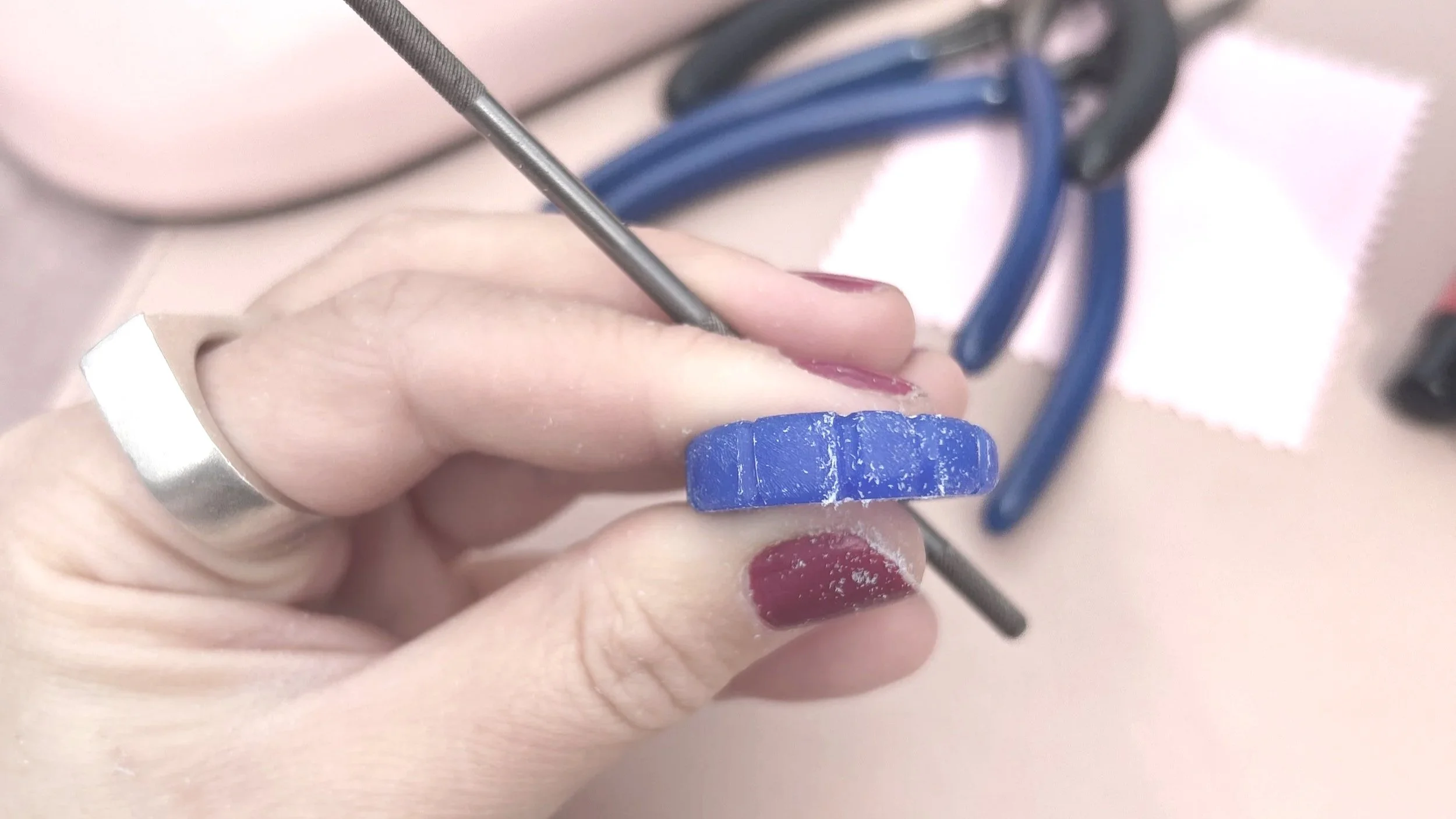 A person with red nail polish holding a small blue object with a white line, while working with a precision tool, with glasses and a cloth in the background.