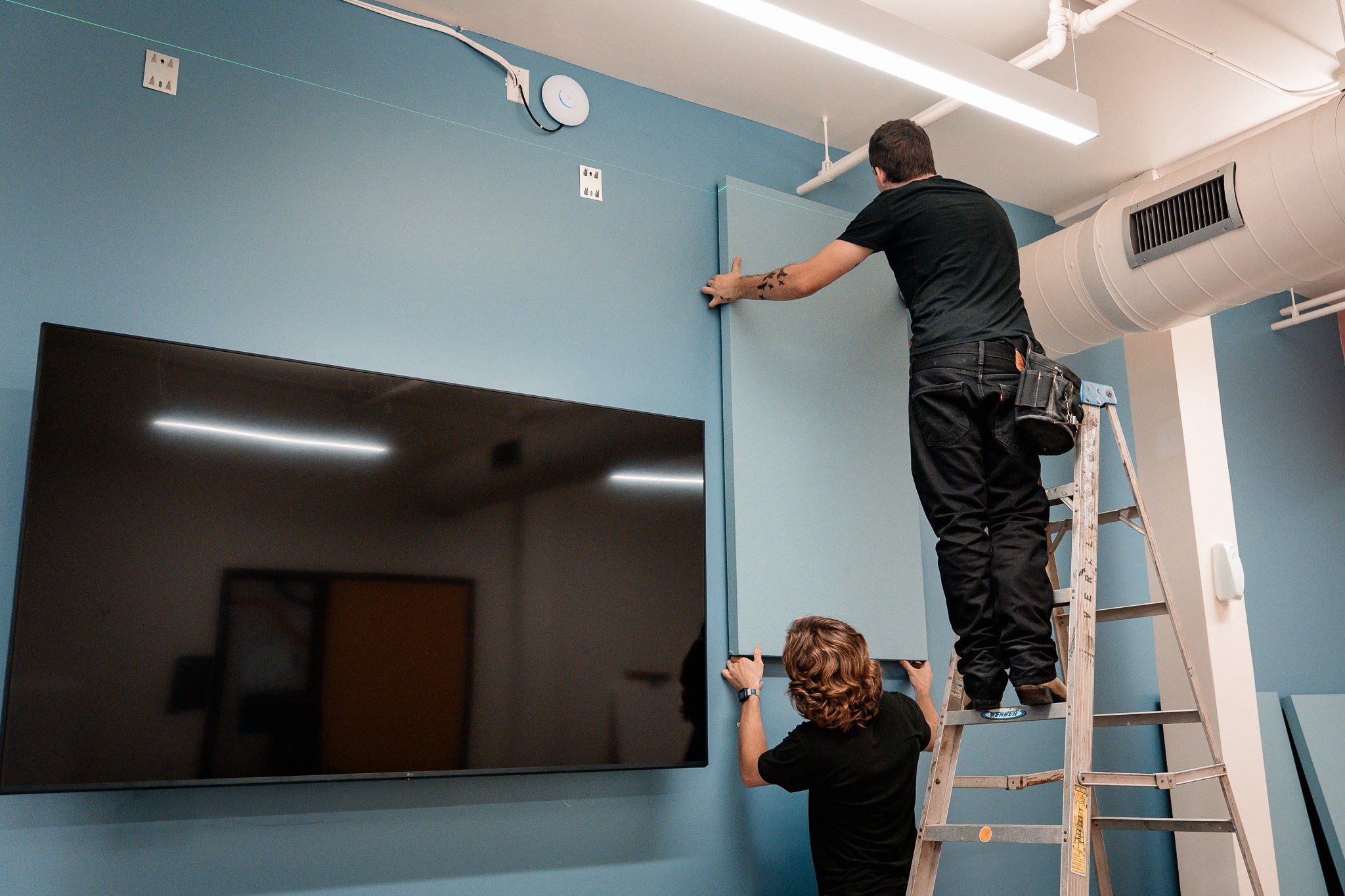 Two men installing a large floating panel on a blue wall near a mounted television in a room with ceiling vents and lighting.
