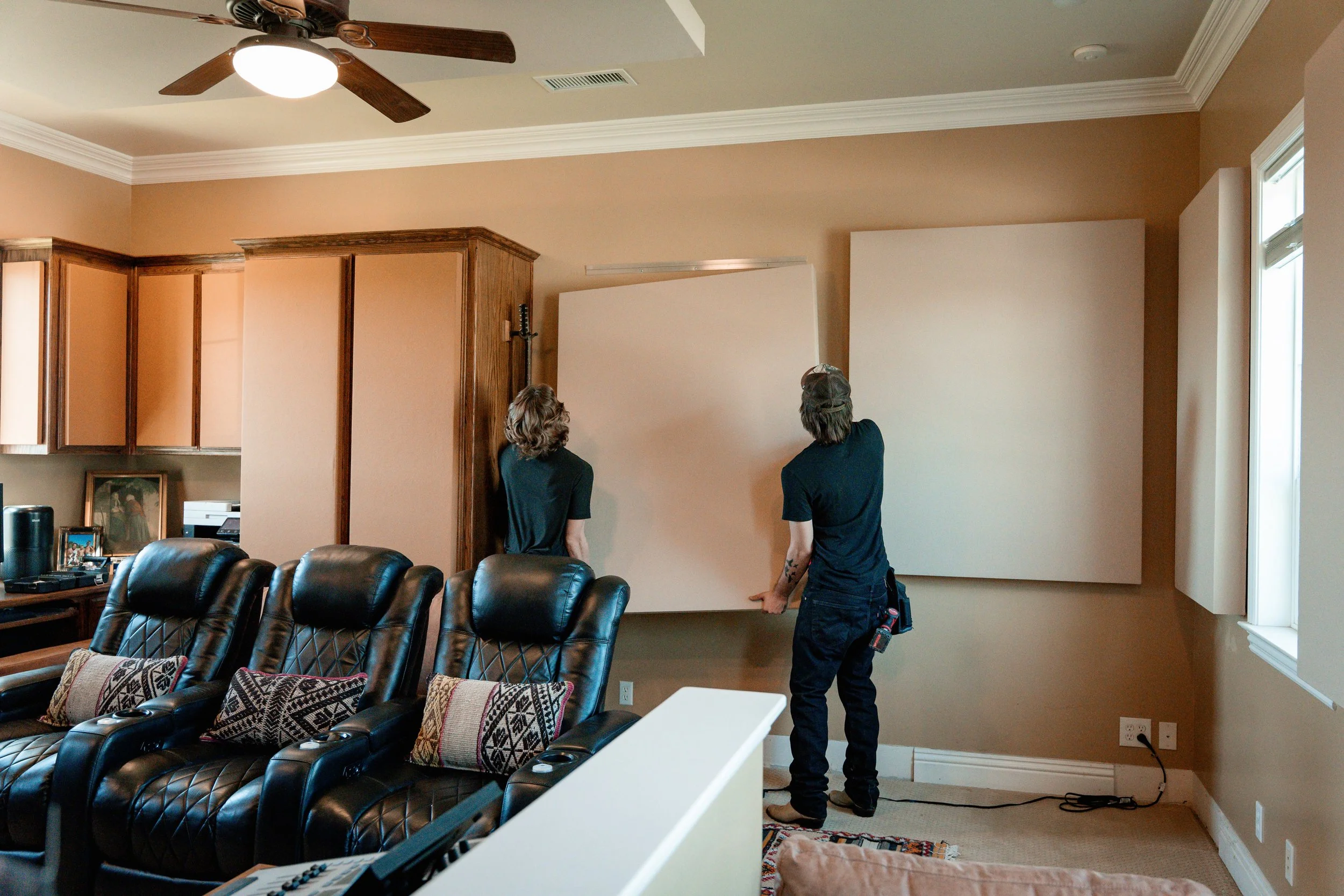 Two workers installing large white soundproof panels on a beige wall in a living room with leather chairs and wooden cabinets.