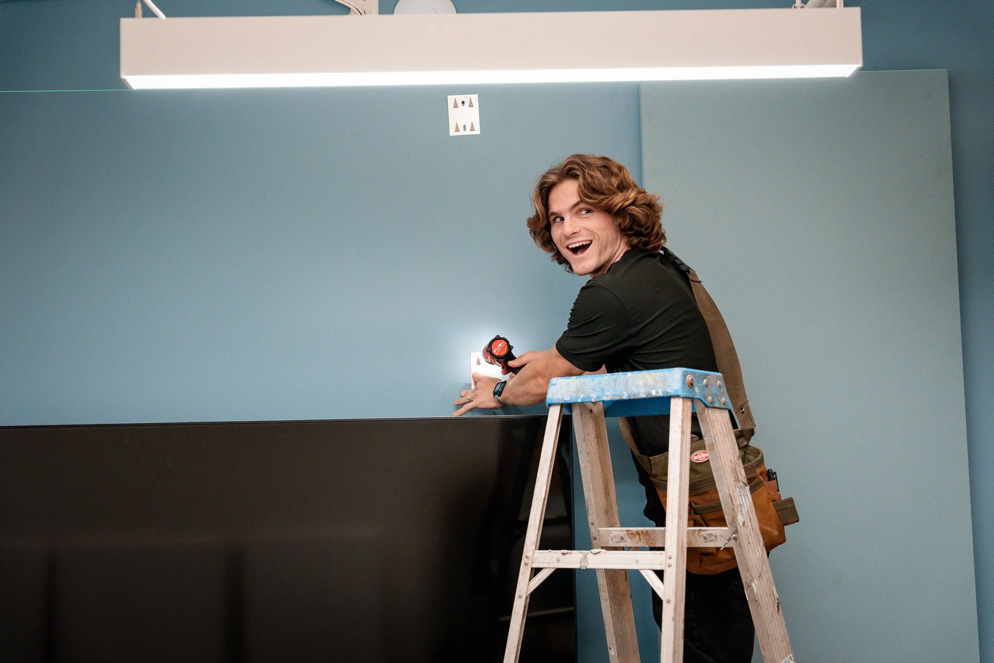 A man with curly hair standing on a step ladder, working on a wall light fixture with a flashlight.