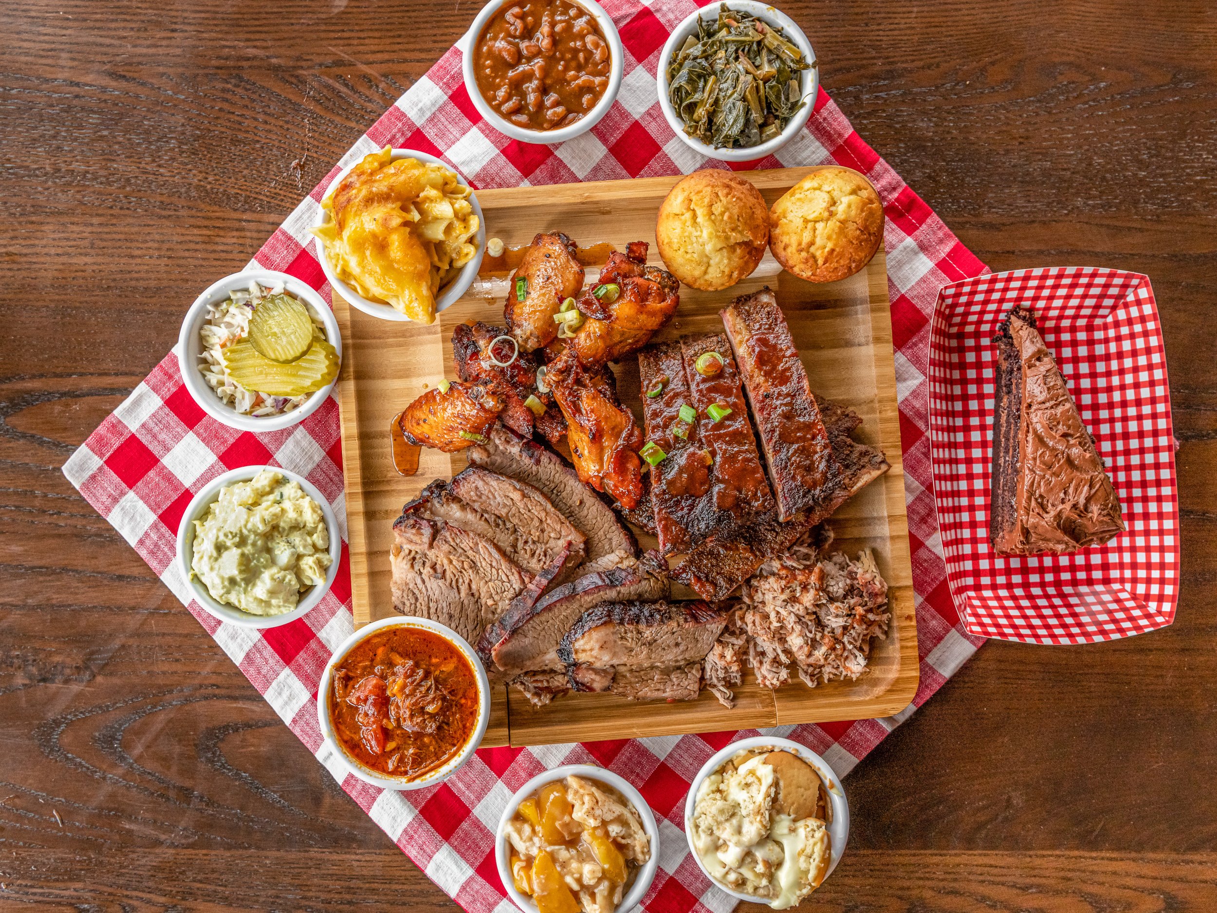 A large wooden platter with various barbecue meats, two cornbread muffins, and multiple side dishes including baked beans, collard greens, pickles, potato salad, and mashed potatoes. A slice of chocolate cake is on a red checked plate to the right, with a red and white checkered tablecloth underneath.