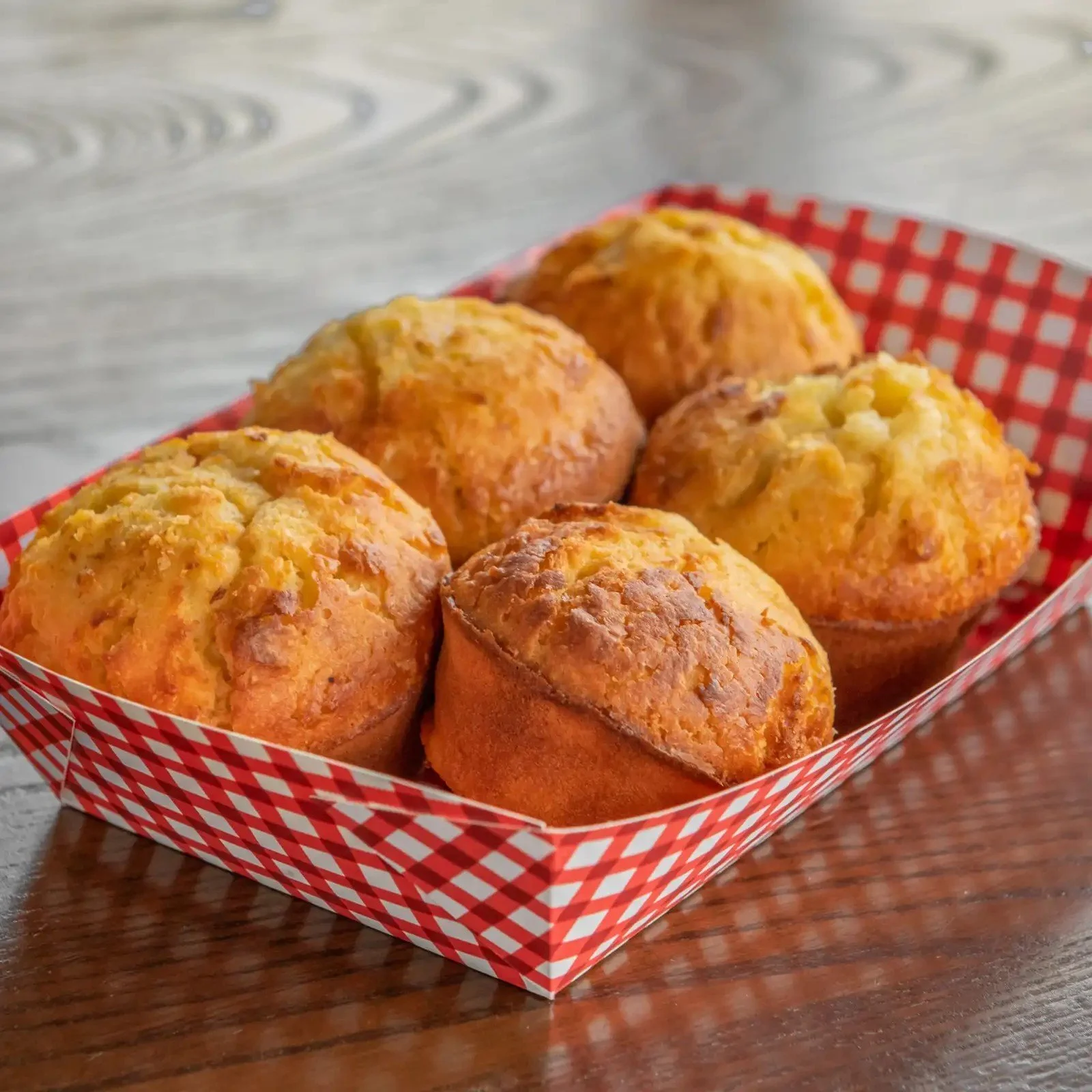 Six freshly baked muffins in a red and white checkered paper tray on a wooden table.