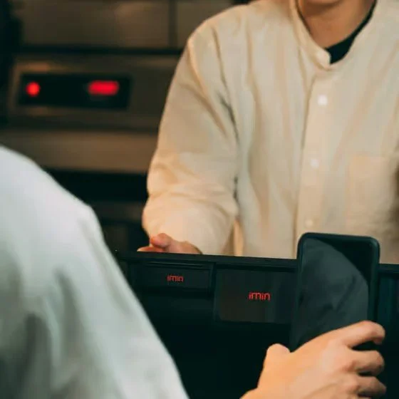 Person in a white shirt handing over a phone to another person in a white shirt at a counter, with a kitchen or restaurant setting in the background.