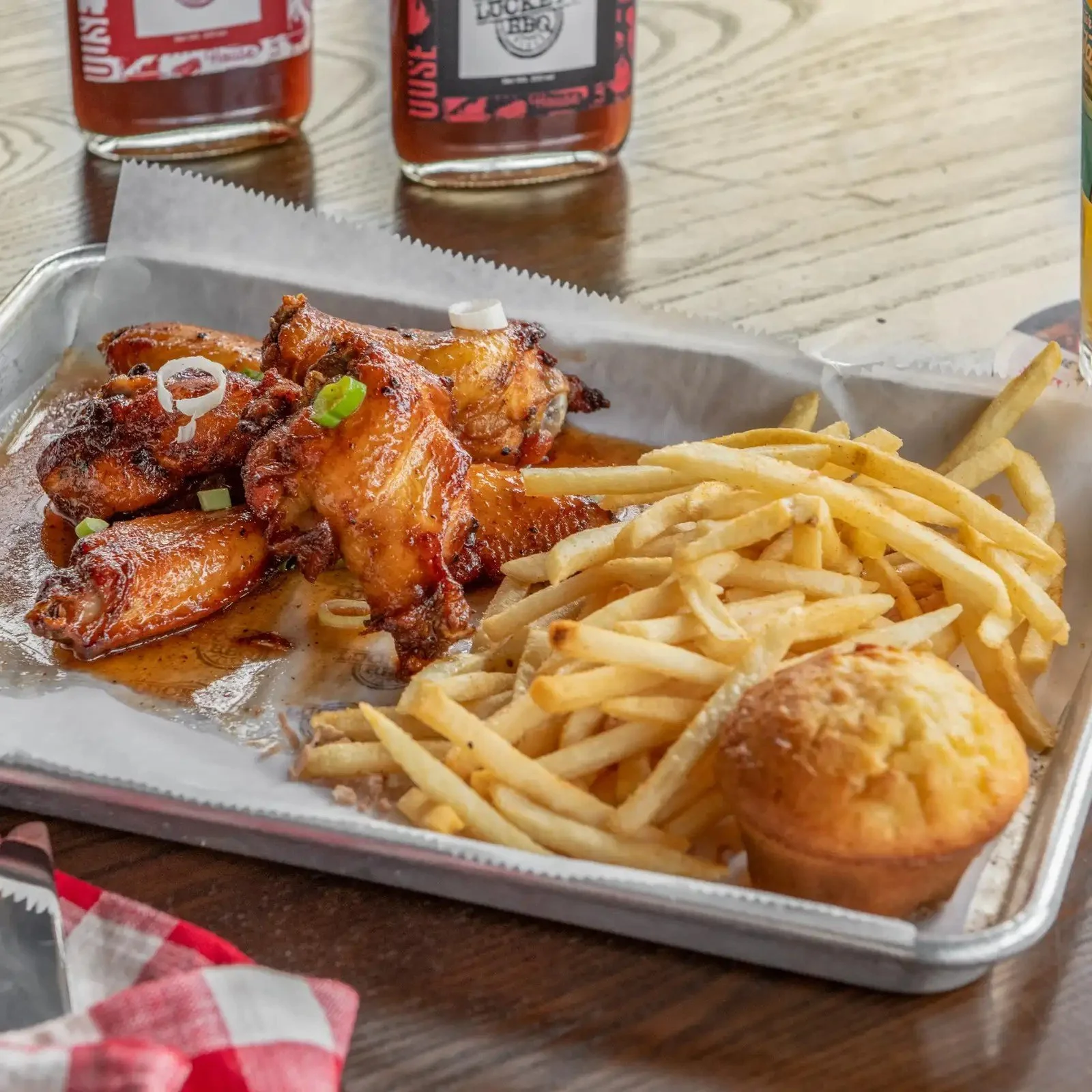 Tray with chicken wings coated in sauce, French fries, and a cornbread muffin, with bottles of hot sauce in the background.