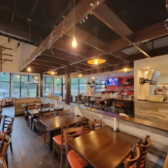 Interior of a modern restaurant with wooden tables and chairs, large windows, and warm lighting.