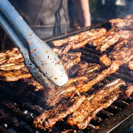 Close-up of a grill with racks of ribs cooking, someone using tongs to handle the meat.